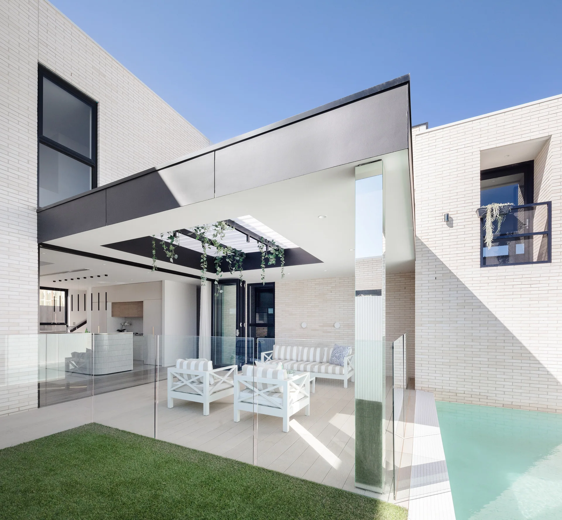 A modern outdoor patio features striped white lounge furniture under a black metal canopy, situated next to a swimming pool and white brick walls.