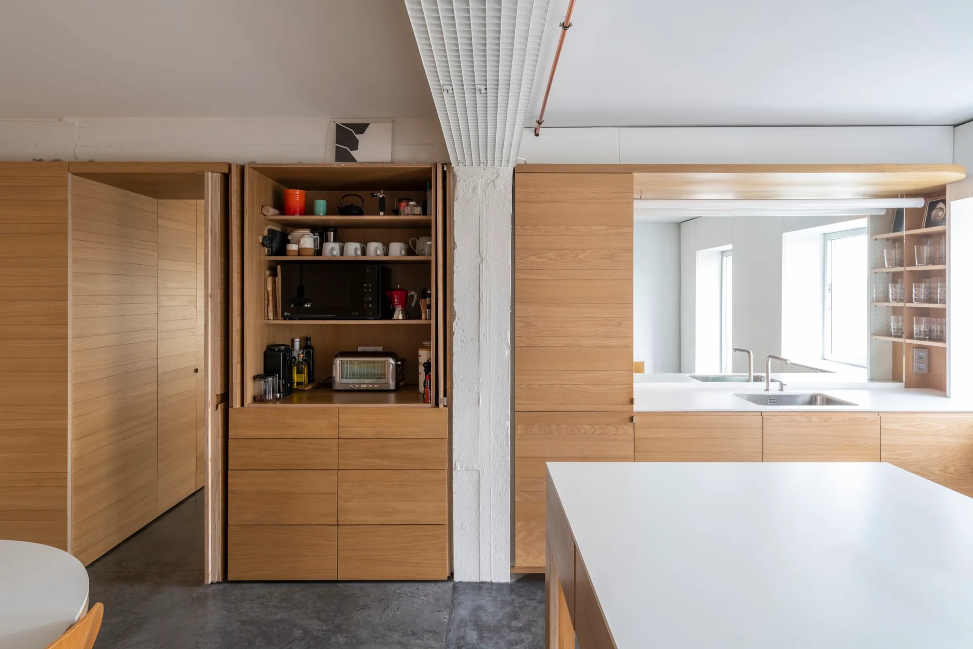 A modern kitchen features light oak cabinetry with an open appliance garage, white countertops, a central white column, and concrete floors.