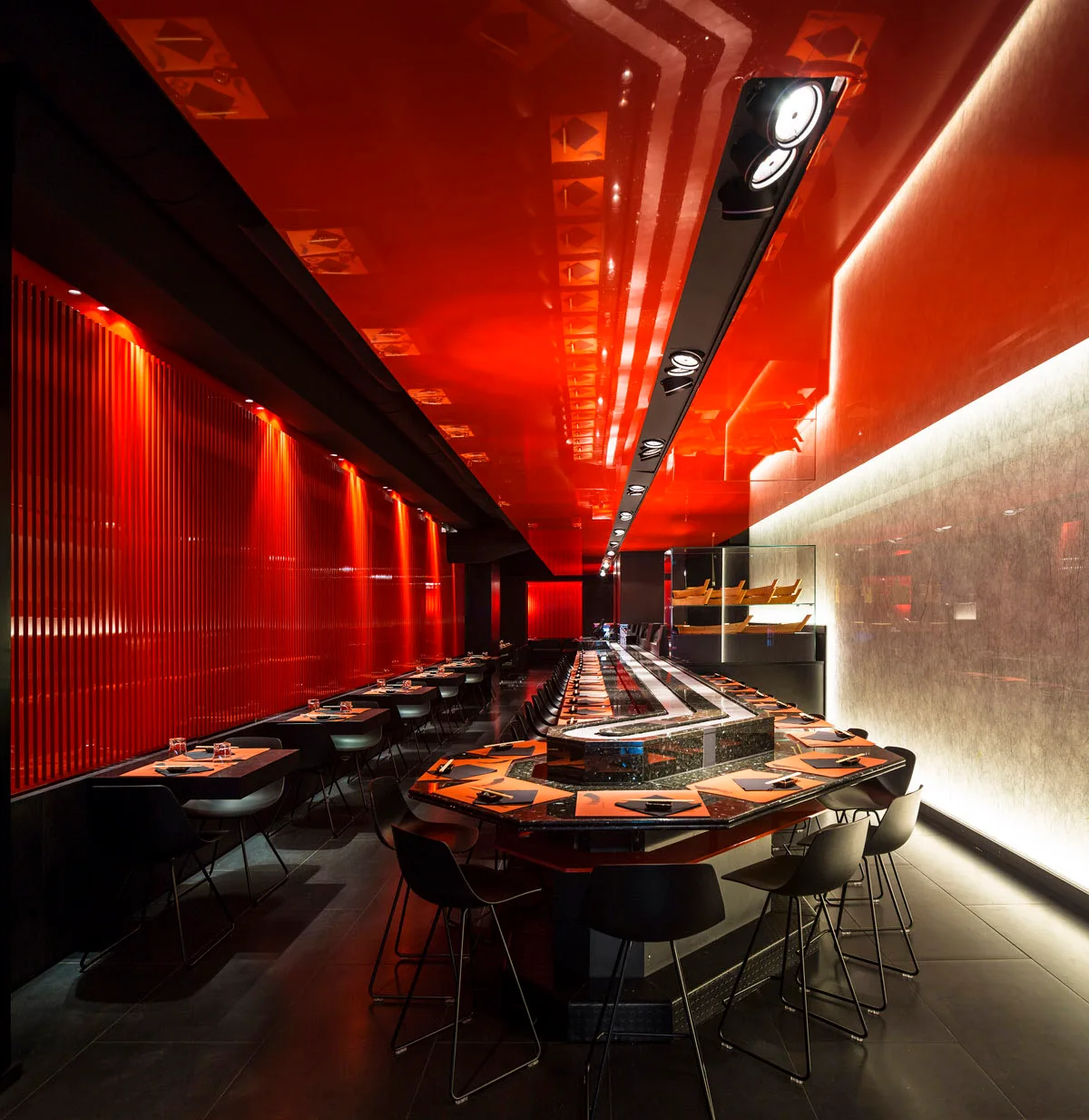 A modern restaurant interior featuring a central dark stone-like bar counter, black stools, a reflective red ceiling, and red vertical slatted walls.