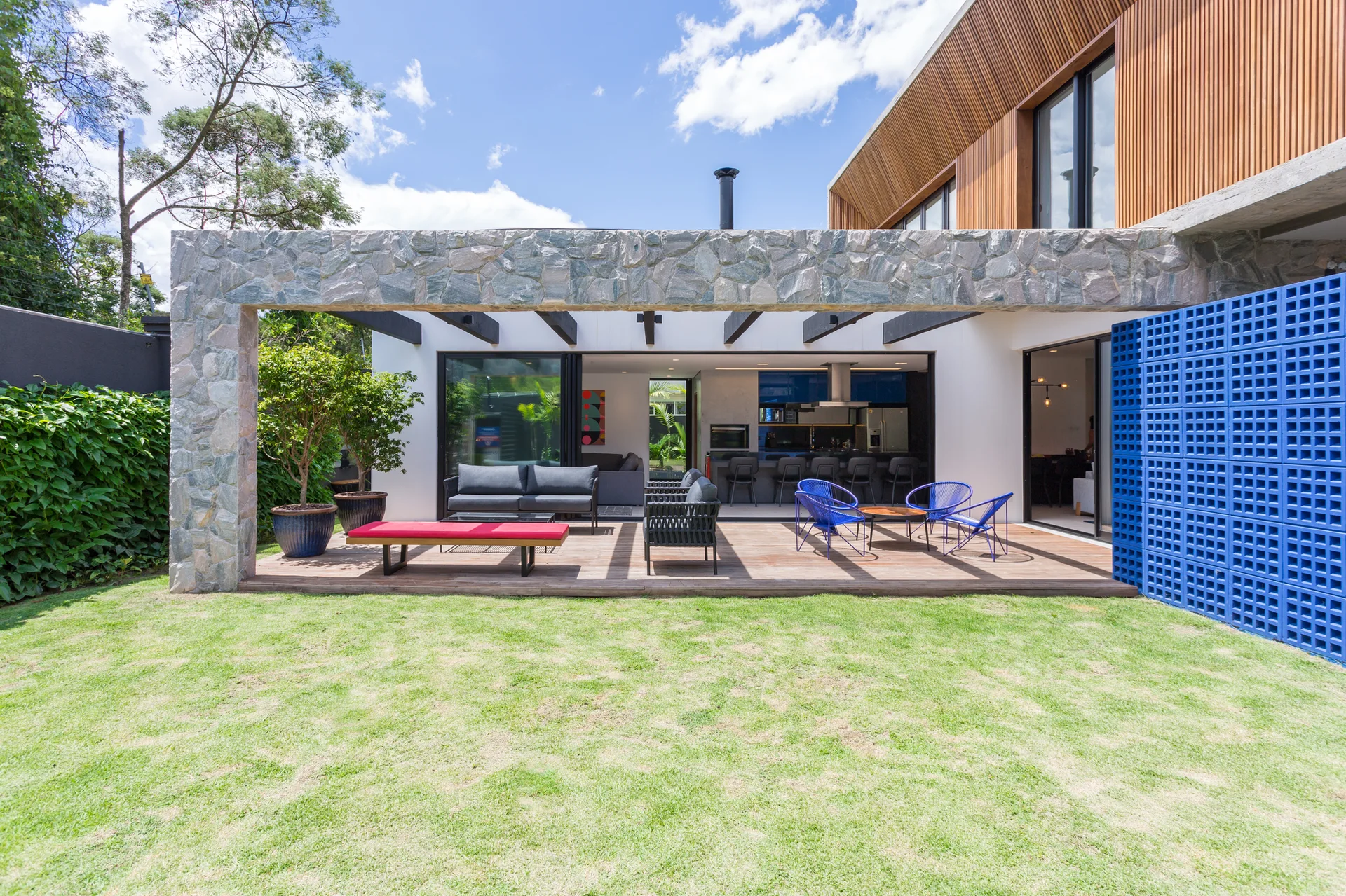 A modern backyard wood deck with colorful outdoor seating, a massive rustic stone overhead frame, and a cobalt blue breeze block wall.