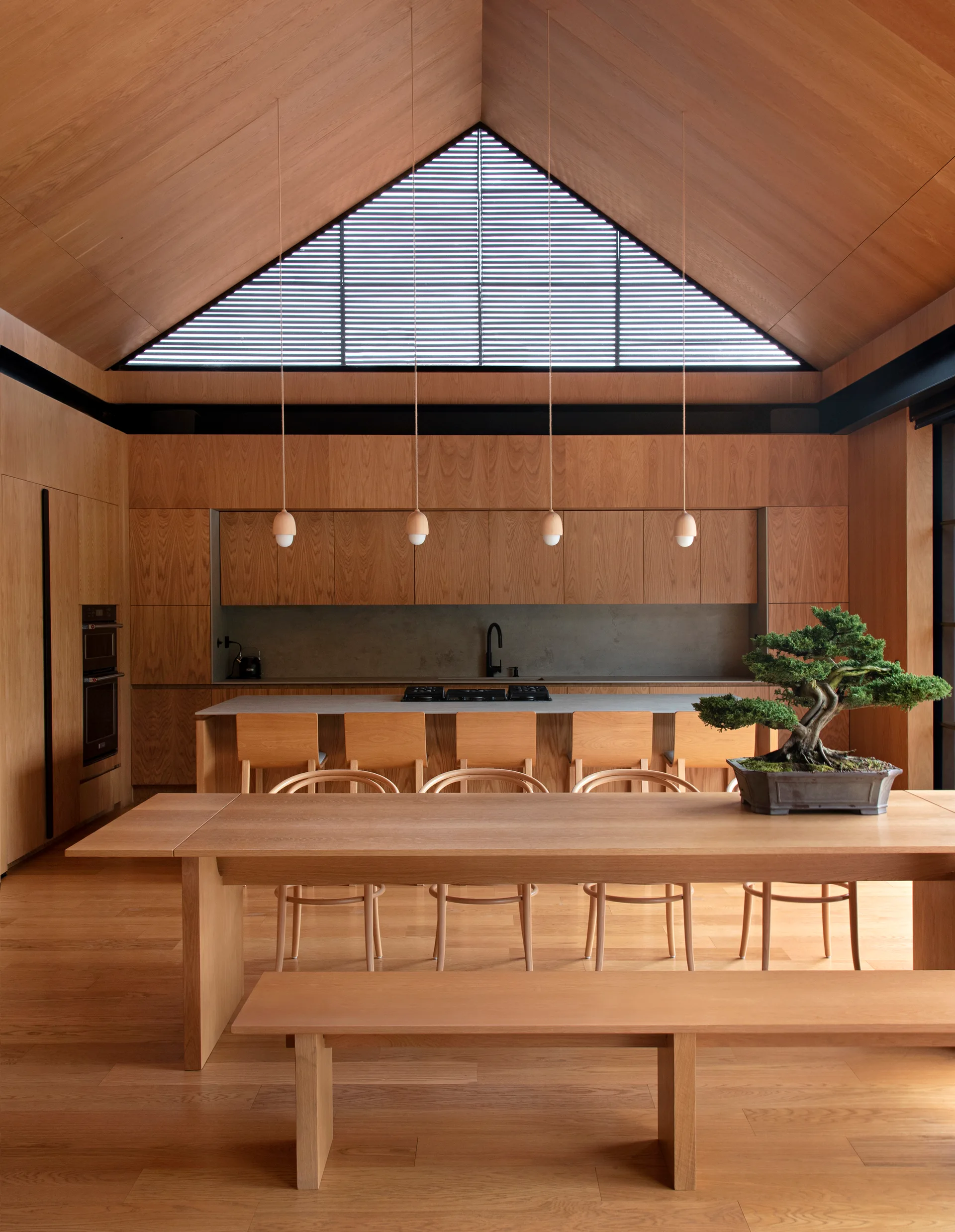 A minimalist kitchen with a vaulted wood-clad ceiling, a triangular high window, a concrete-topped island, and a long wooden dining table with a bonsai tree.