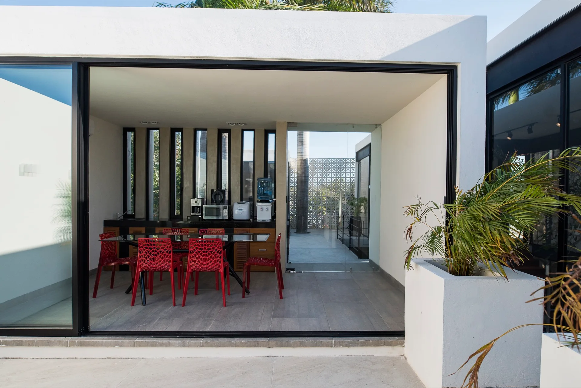 A modern covered patio with an outdoor kitchen, a glass-top dining table, and six vibrant red lattice chairs.