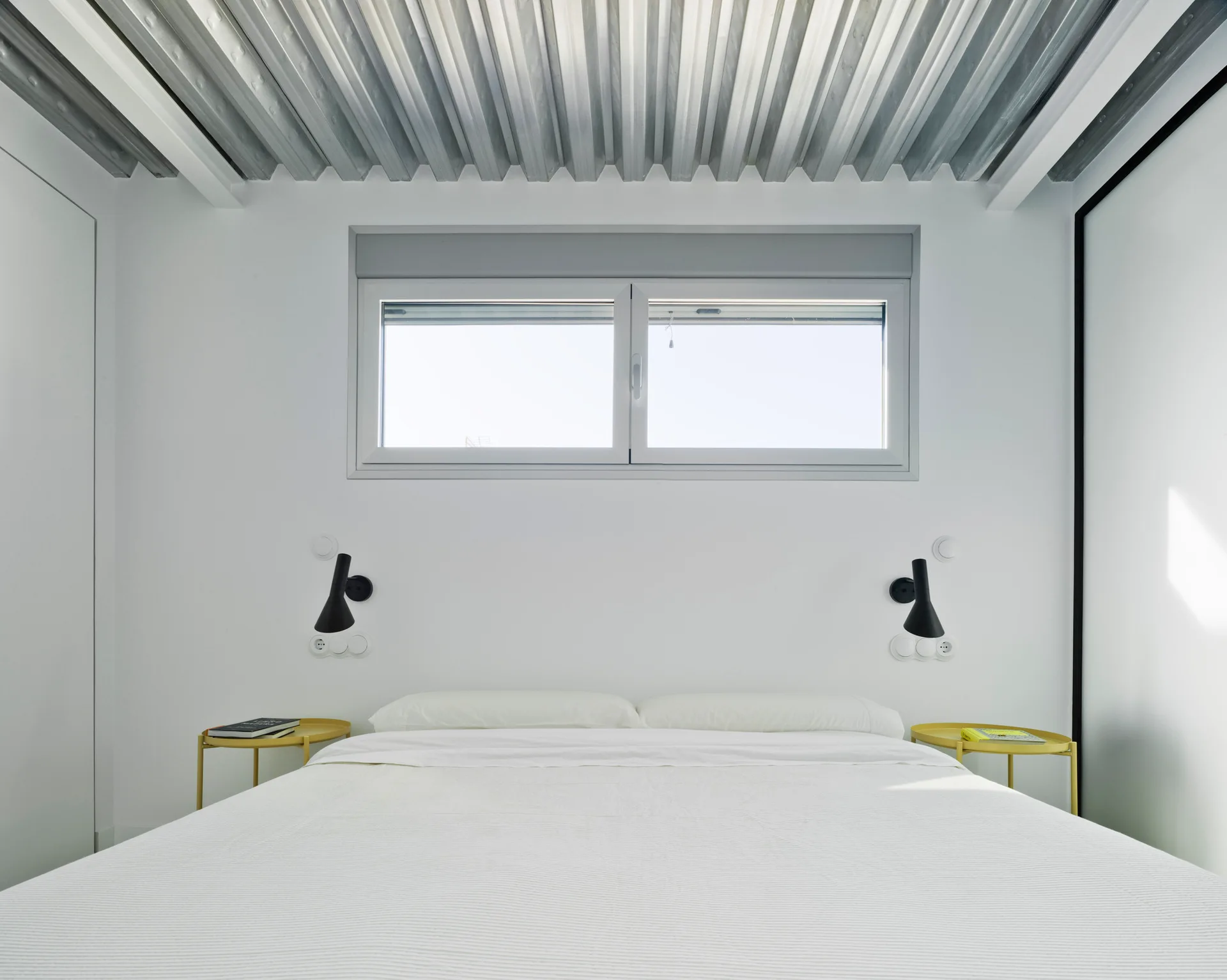 Minimalist bedroom with a white bed under a horizontal window, flanked by yellow metal side tables and black wall lamps beneath a corrugated metal ceiling.