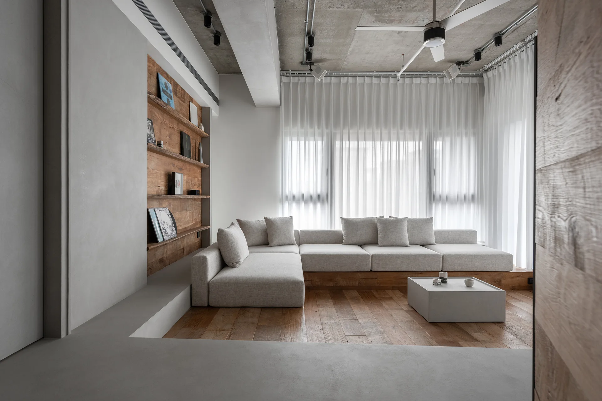 A minimalist living room with a beige sectional sofa on a low wooden platform, recessed wood shelves, and an exposed concrete ceiling.