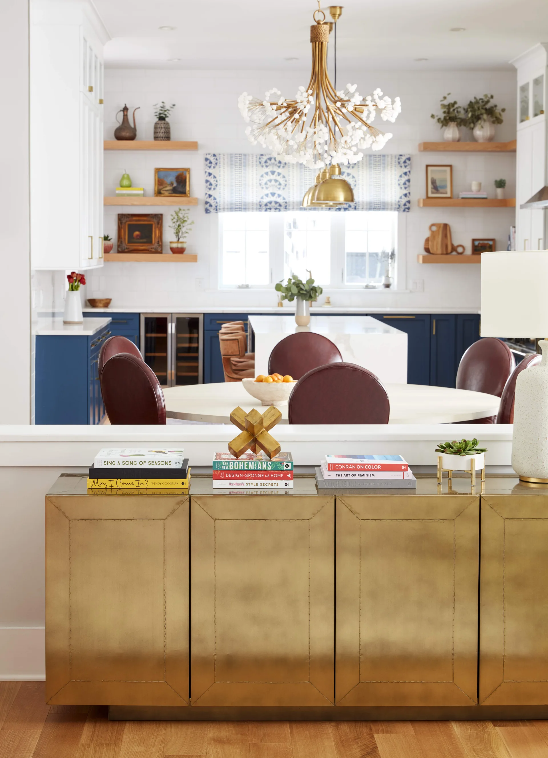 A dining room features a textured brass sideboard in the foreground, circular red leather chairs around a table, and a blue and white kitchen behind.