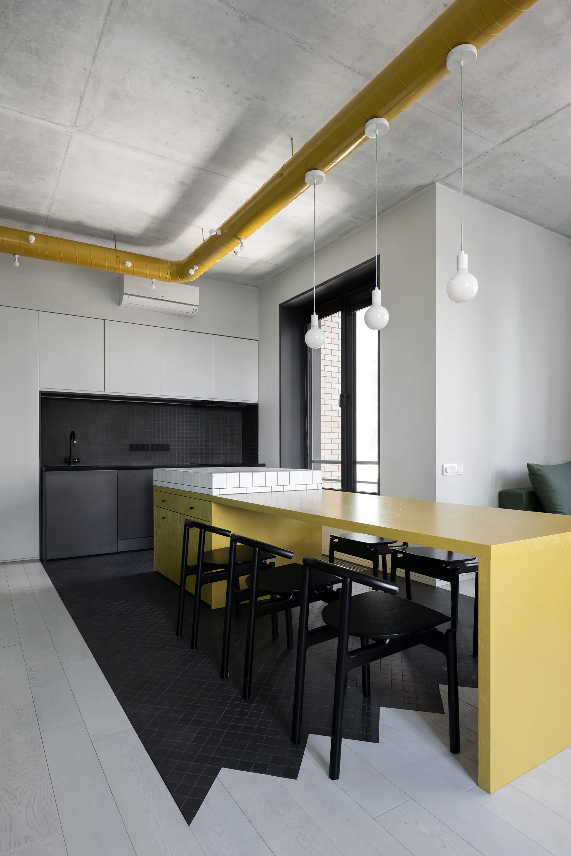 Modern industrial kitchen featuring a long yellow dining island, matching yellow ceiling ductwork, black chairs, and a raw concrete ceiling.