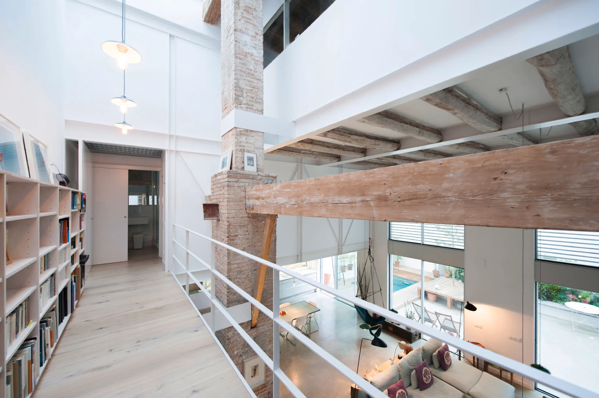 A mezzanine landing with light wood floors and a white bookshelf overlooks a double-height living room with exposed brick and rustic ceiling beams.
