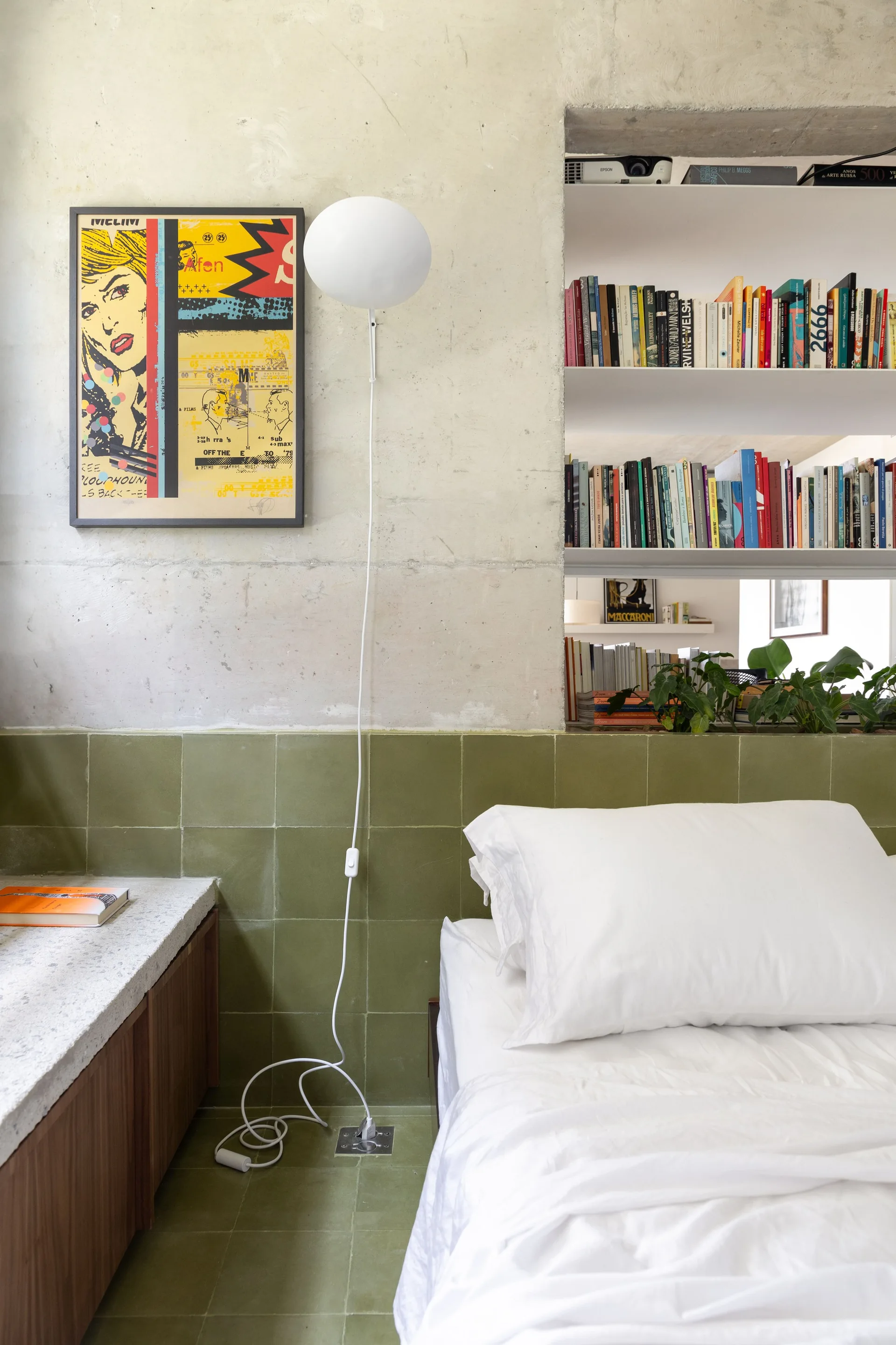 A minimalist bedroom features white bedding next to an olive green tiled wall, raw concrete, and a built-in bookshelf niche.
