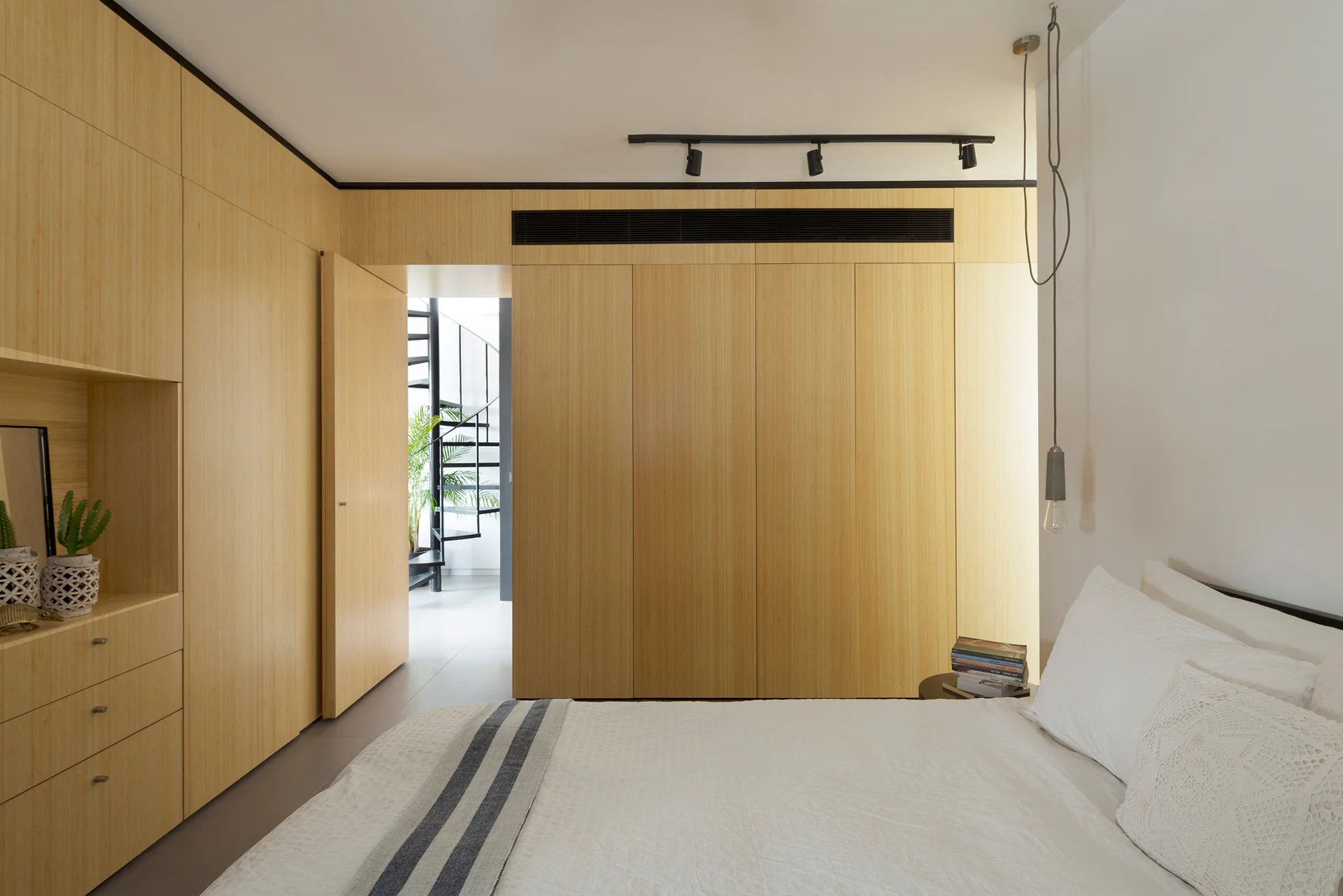 A modern bedroom with white bedding, custom floor-to-ceiling light wood cabinetry, track lighting, and a view of a spiral staircase.
