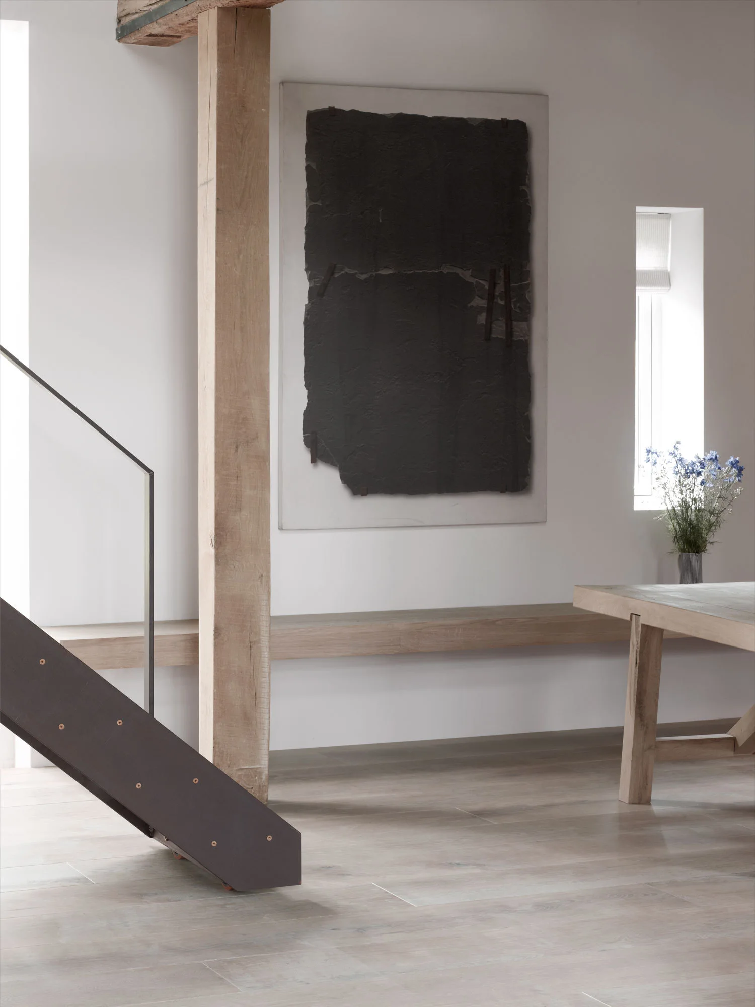 A minimalist room featuring an exposed vertical wood post, a dark textured art piece on a white wall, a floating wooden bench, and a wooden table.