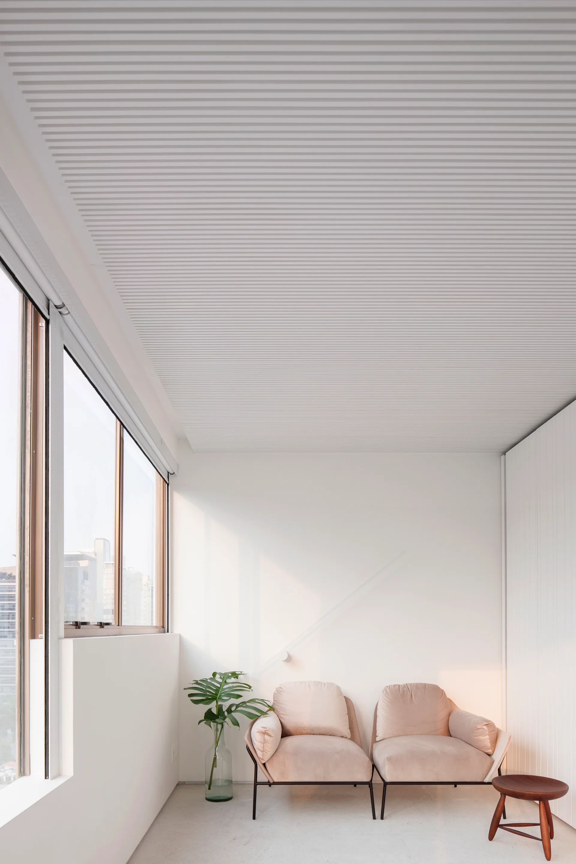 Two beige upholstered armchairs sit side-by-side in a bright, minimalist room featuring a white slatted ceiling and a large window.
