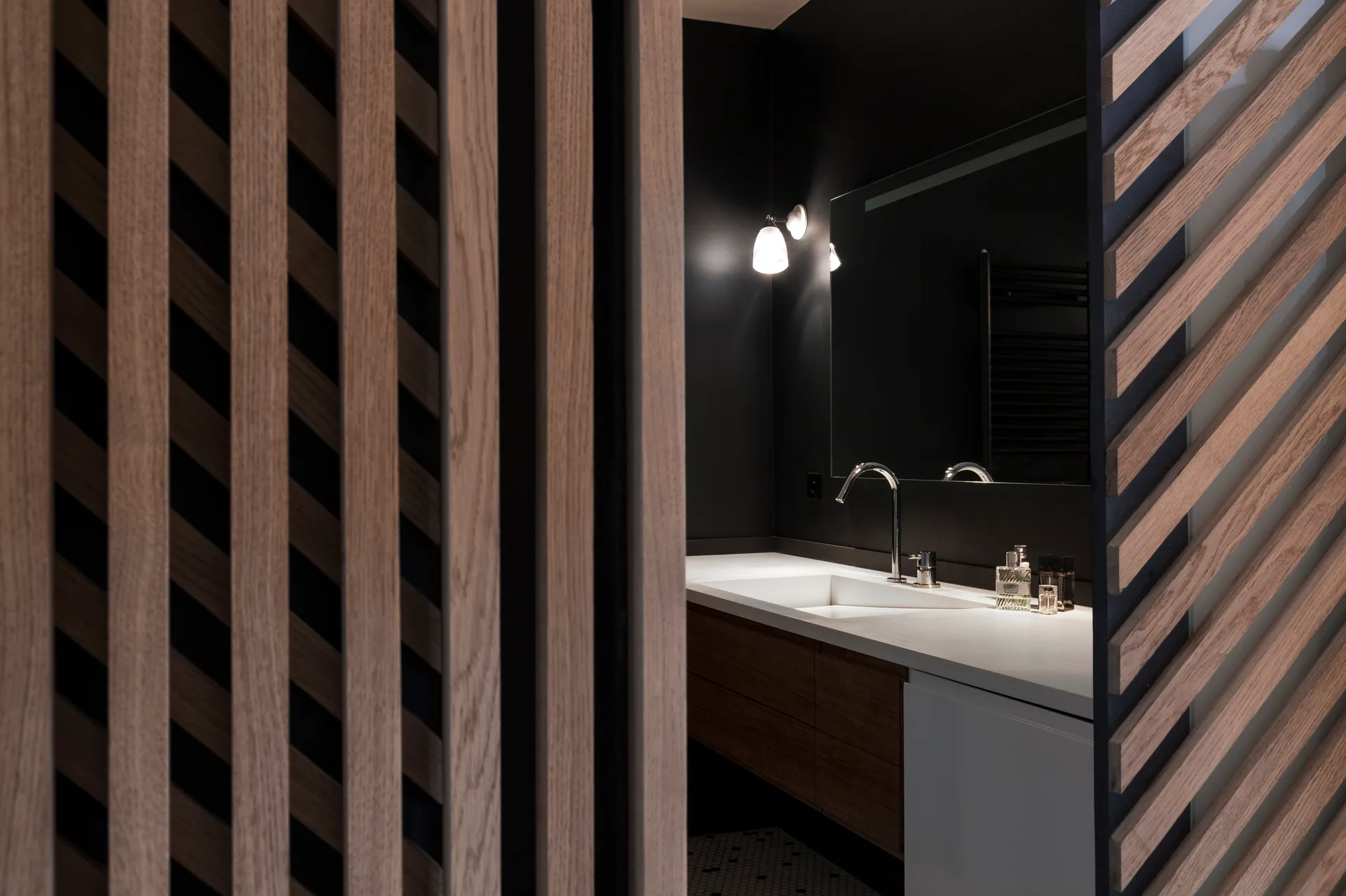 A modern bathroom vanity with a white integrated sink is visible through a diagonal wooden slat screen against a dark wall.