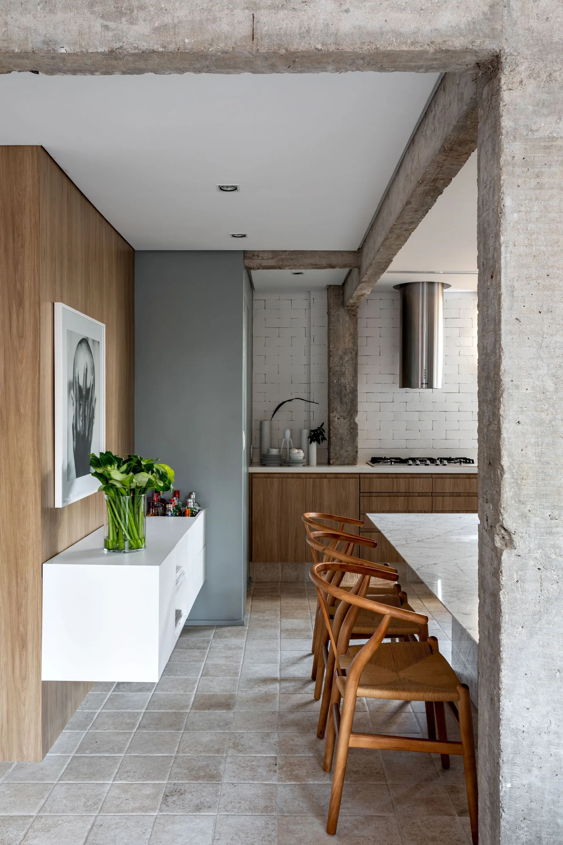 An industrial modern kitchen featuring exposed concrete columns, natural wood cabinets, woven wooden dining chairs at a marble island, and stone tile flooring.