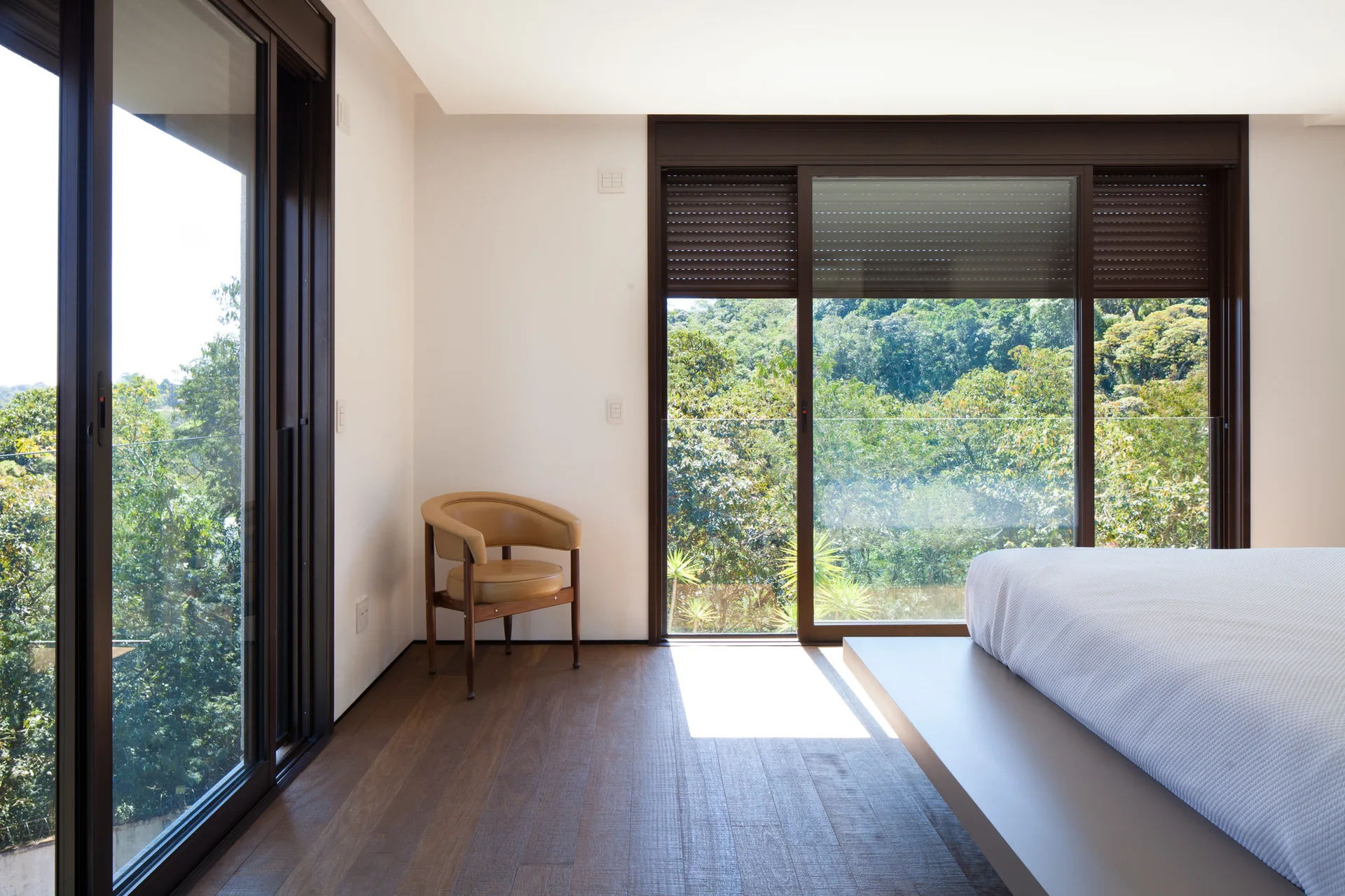 A minimalist bedroom featuring dark wood floors, a low-profile white bed, a tan leather armchair, and large sliding glass doors overlooking a lush forest.