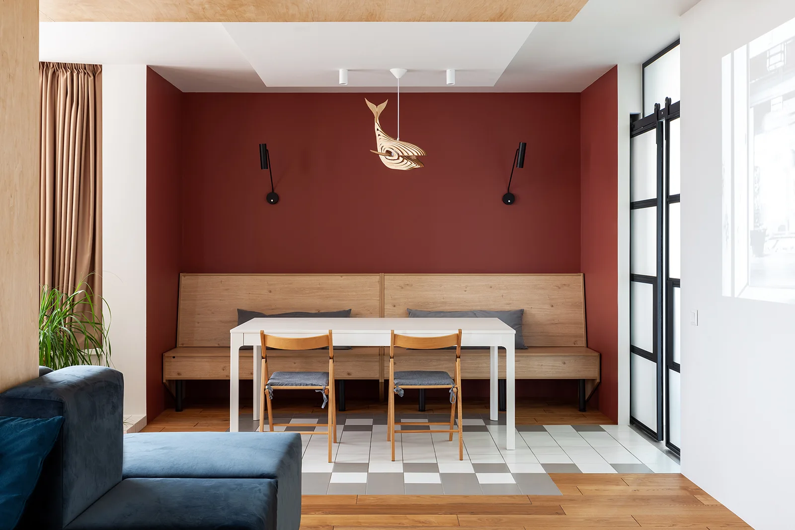 A modern dining area with a white table, a wooden bench against a dark red accent wall, and a checkerboard floor tile inlay.