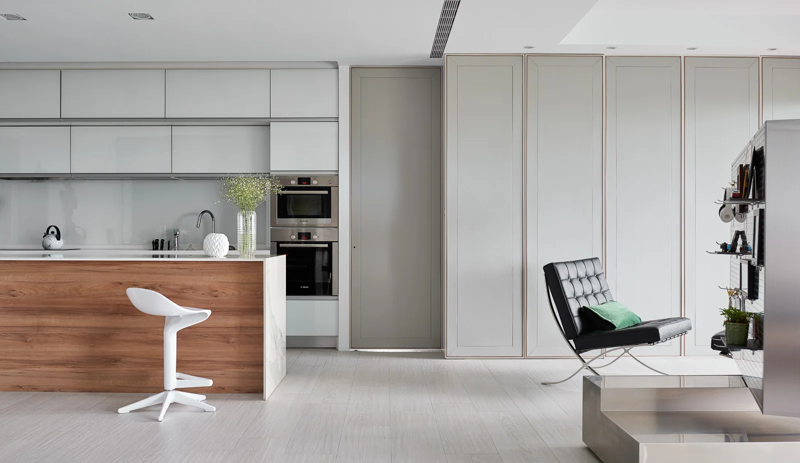 A minimalist kitchen features a wood-grain island with a white bar stool, white cabinetry, integrated appliances, and a black tufted leather lounge chair.