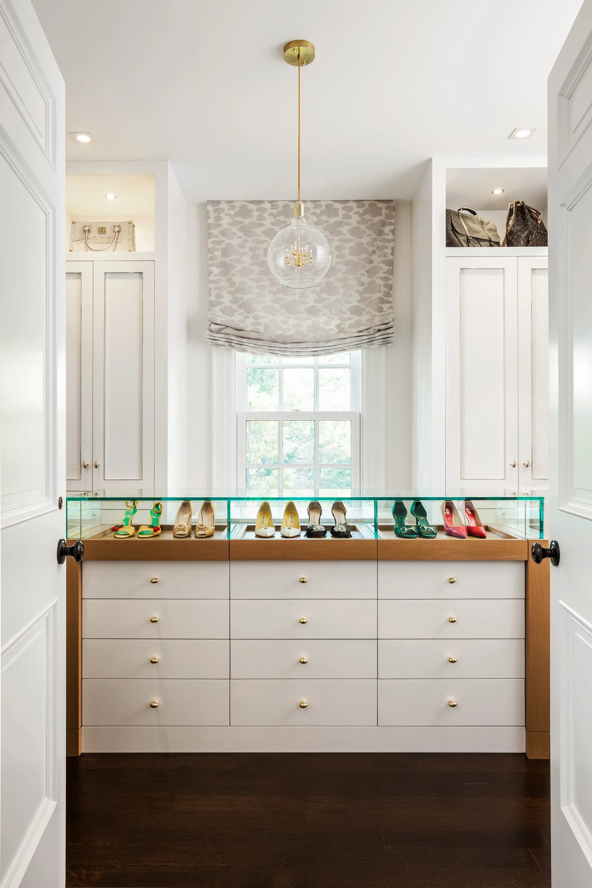 A white dressing room viewed through open double doors features built-in cabinets and a dresser with a glass-topped shoe display case.