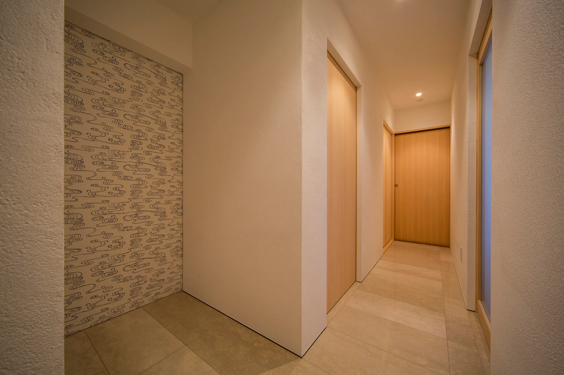 A minimalist hallway features light wood flush doors, a stone tile floor, and a recessed alcove with black and white wavy patterned wallpaper.