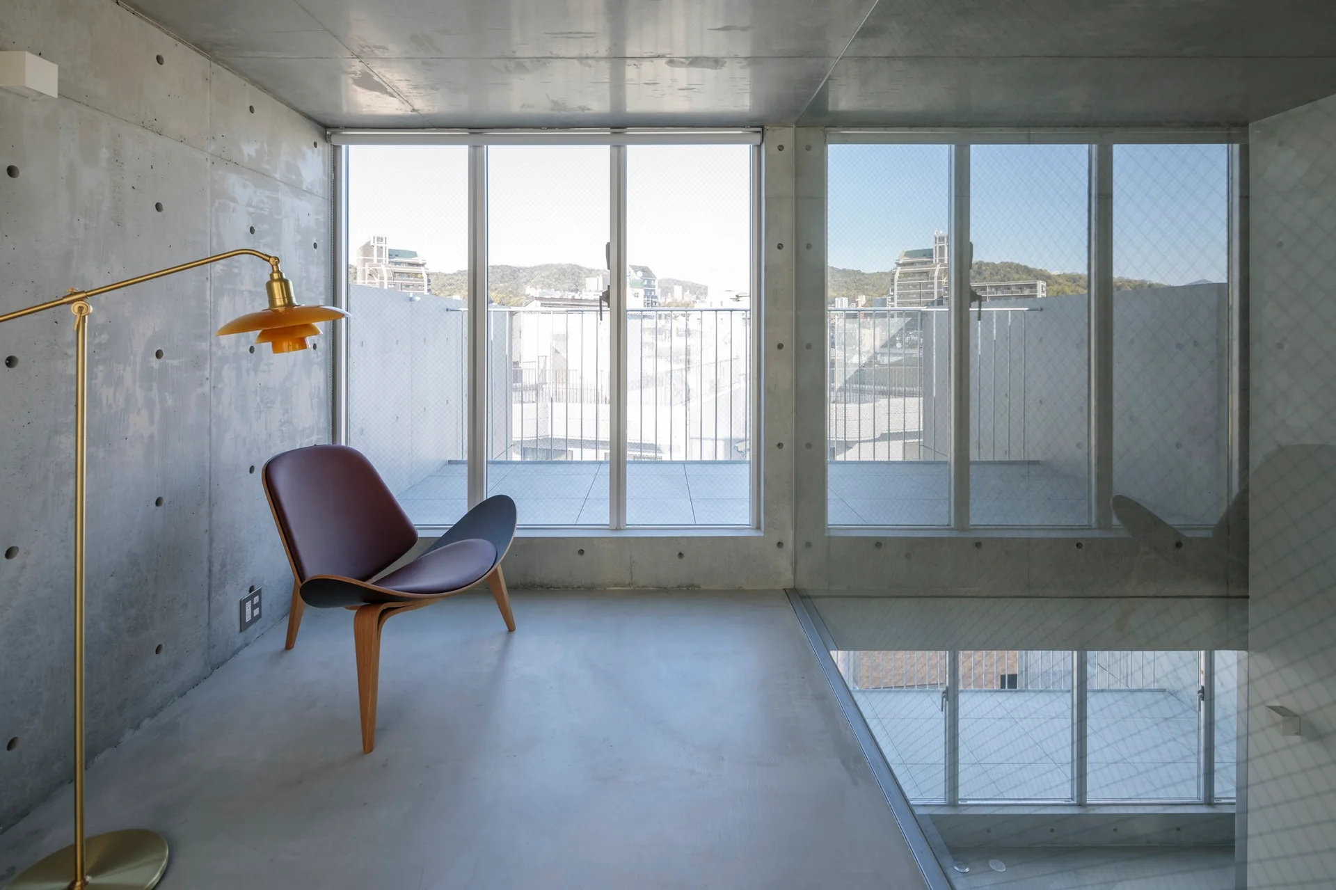 A minimalist room with exposed concrete walls features a curved wooden shell chair and a brass floor lamp in front of large glass doors.