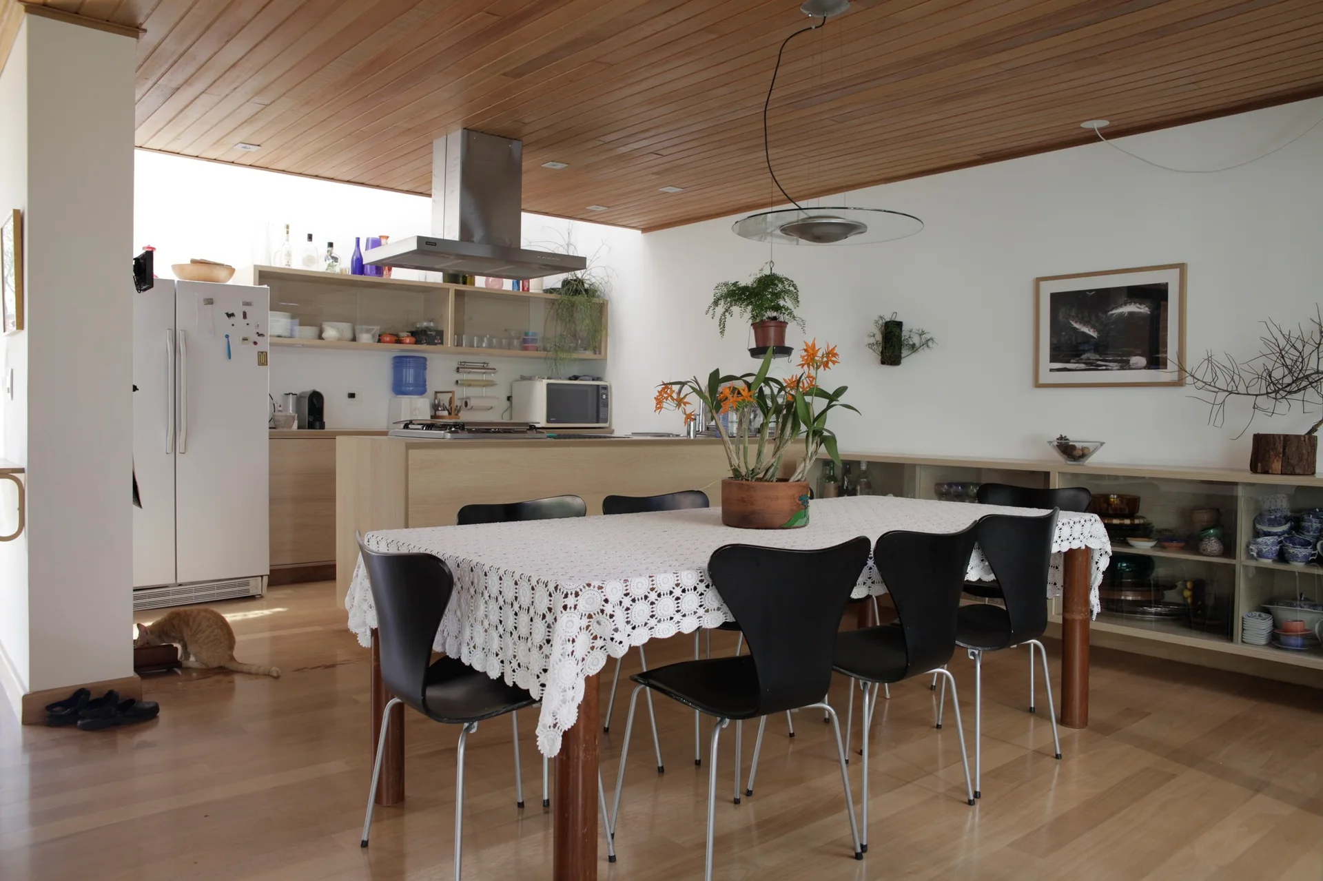 A dining room features a wooden table with a white lace tablecloth and black chairs beneath a slatted wood ceiling next to a kitchen.