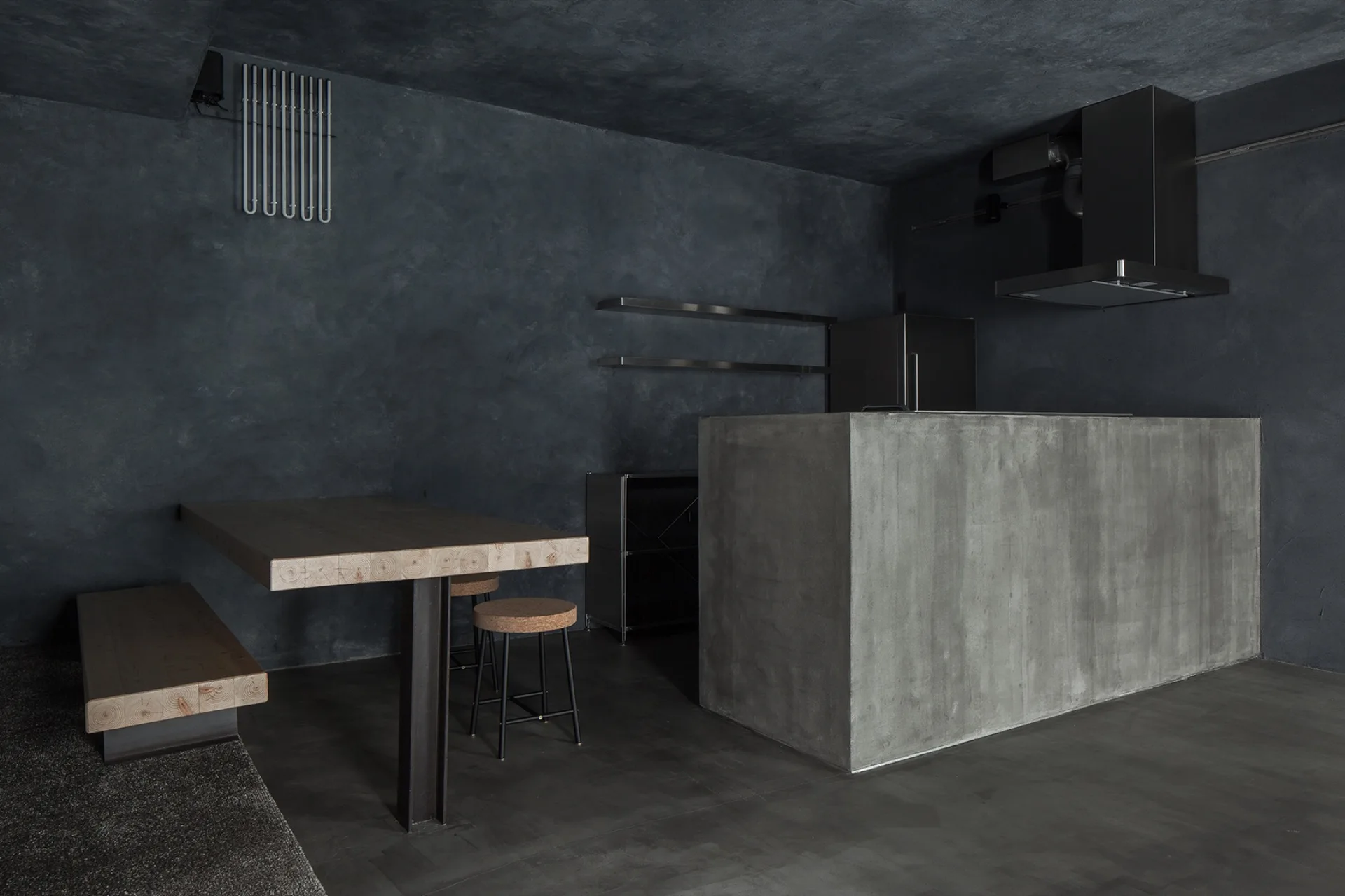 A minimalist dark grey kitchen featuring a massive concrete island, textured charcoal plaster walls, and a light wood dining table with cork stools.