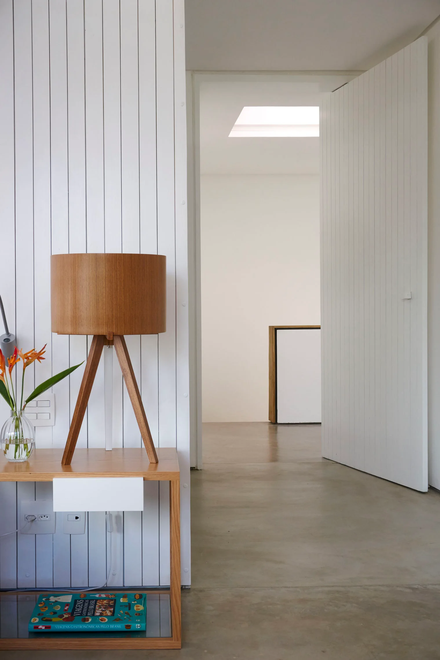 A modern hallway features a light wood side table with a wooden tripod lamp against a white vertical slatted wall and an open paneled door.