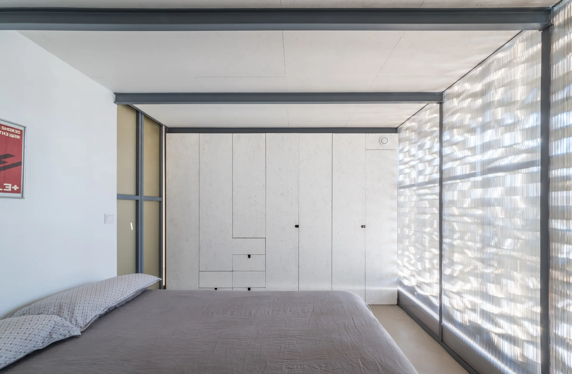 A minimalist bedroom features a grey bed next to a large white built-in wardrobe, dark metal ceiling beams, and a textured translucent window screen.