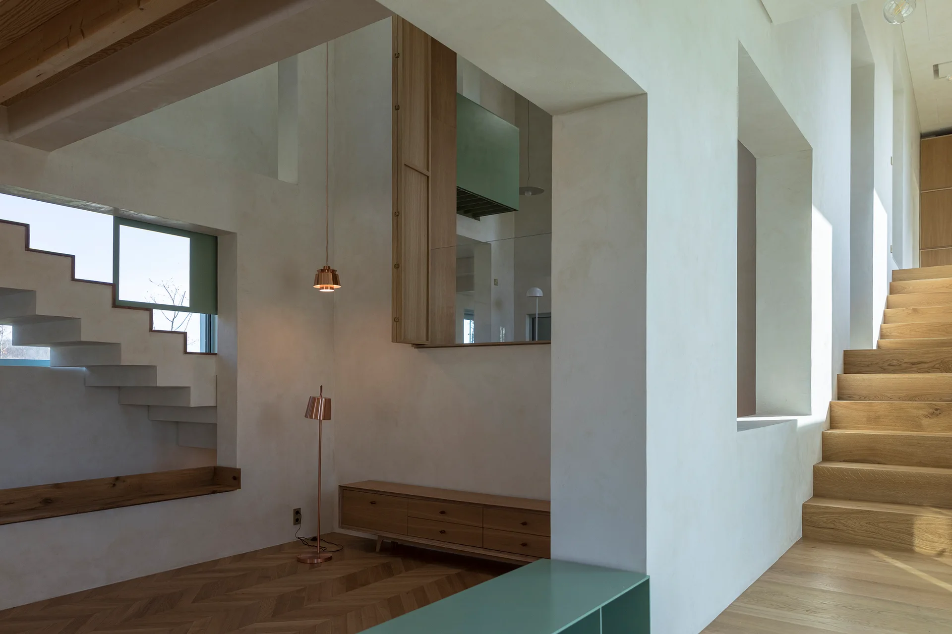 A minimalist interior featuring smooth plaster walls, a wooden staircase, herringbone flooring, and a low oak console cabinet with copper lights.