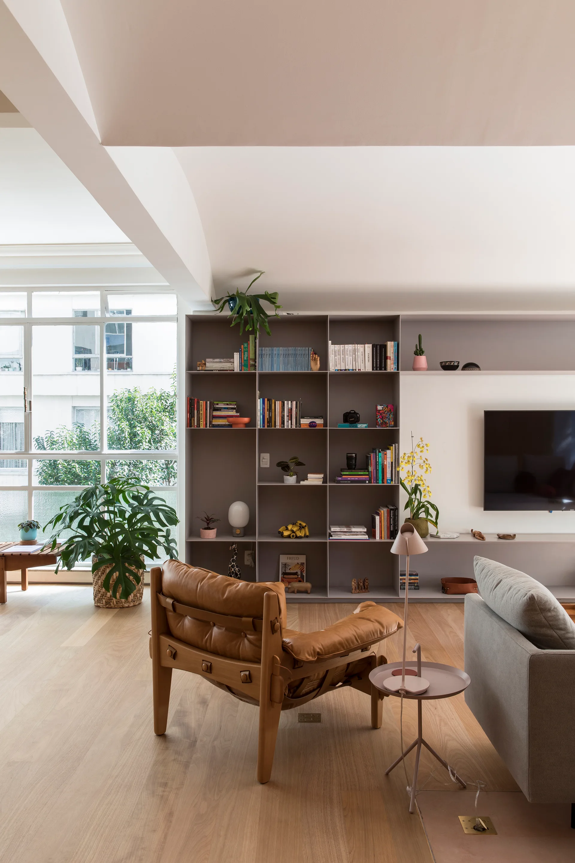 A light-filled living room featuring a brown leather armchair, grey sofa, and a large taupe wall shelving unit with books.