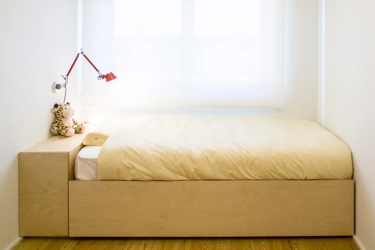 A compact white bedroom alcove features a custom light wood platform bed, a sheer window shade, and a red articulated wall-mounted lamp.