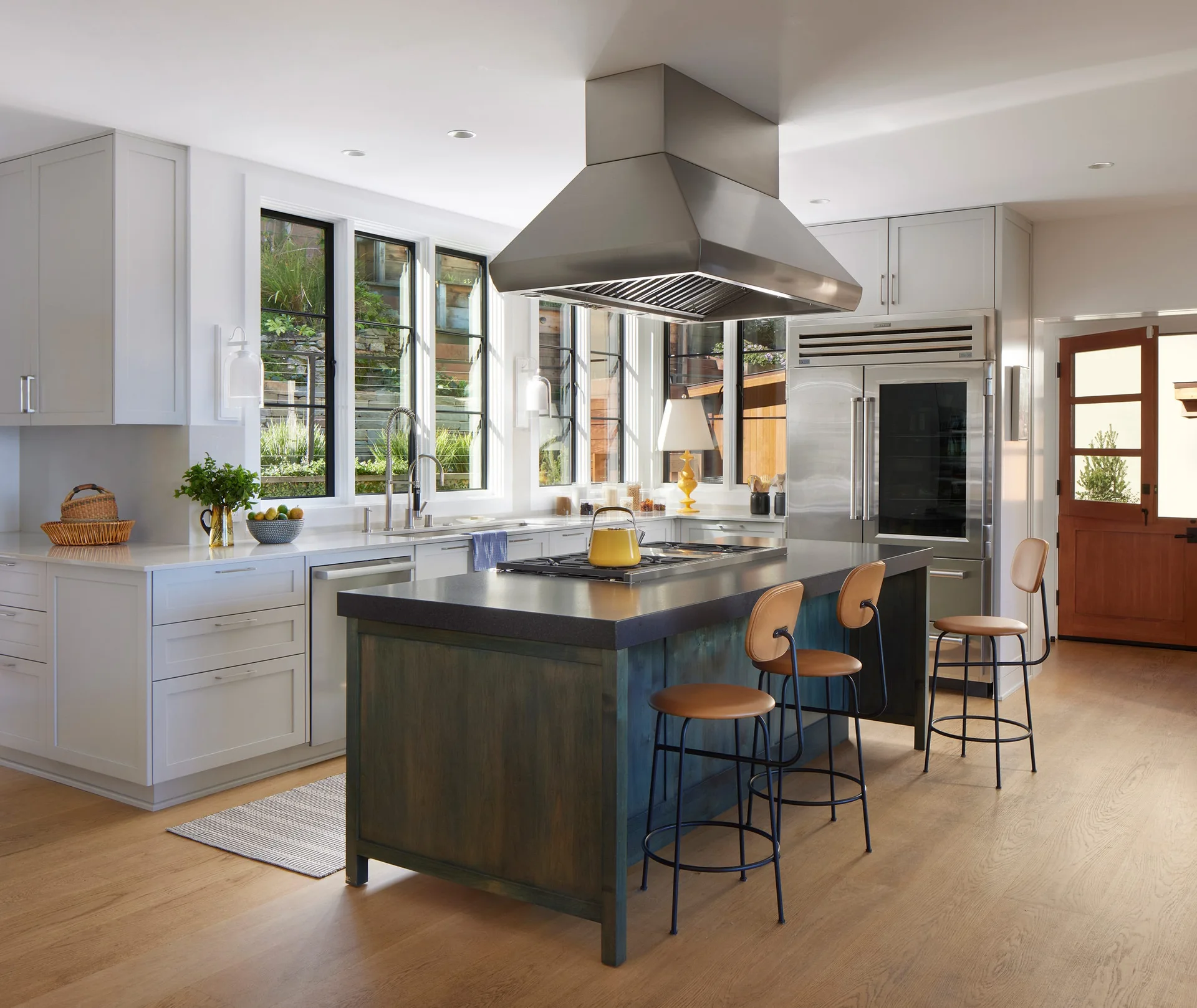 A light gray kitchen features a central dark stained wood island with three leather bar stools, a stainless steel range hood, and black-framed windows.