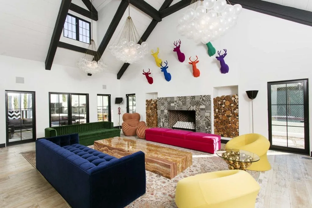 An eclectic living room with a vaulted ceiling, dark wood beams, colorful sofas, yellow armchairs, and a tiled fireplace with stacked wood alcoves.