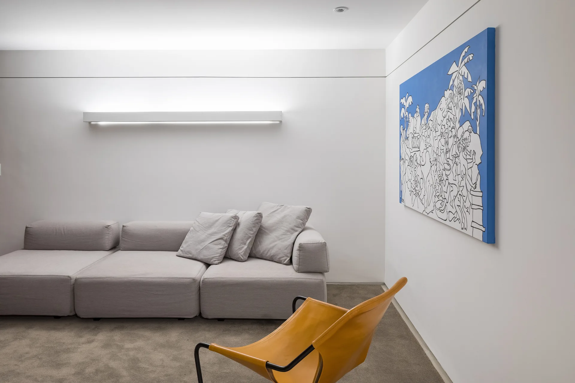 A minimalist living room features a grey modular sofa beneath a long linear wall sconce, next to a large blue painting and a leather chair.