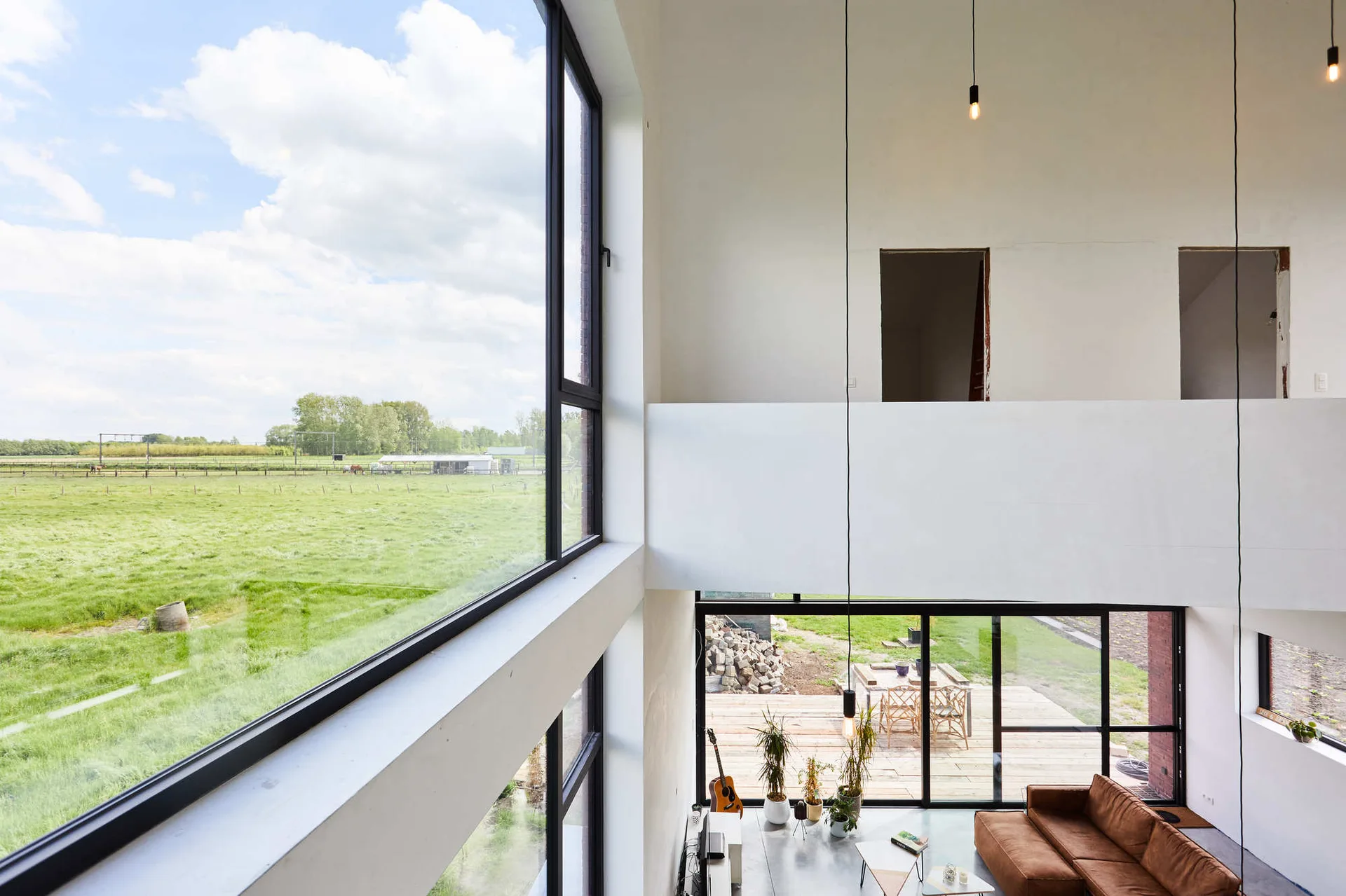 A double-height modern living room features a brown leather sofa, large black-framed windows looking out to a green field, and an upper mezzanine level.