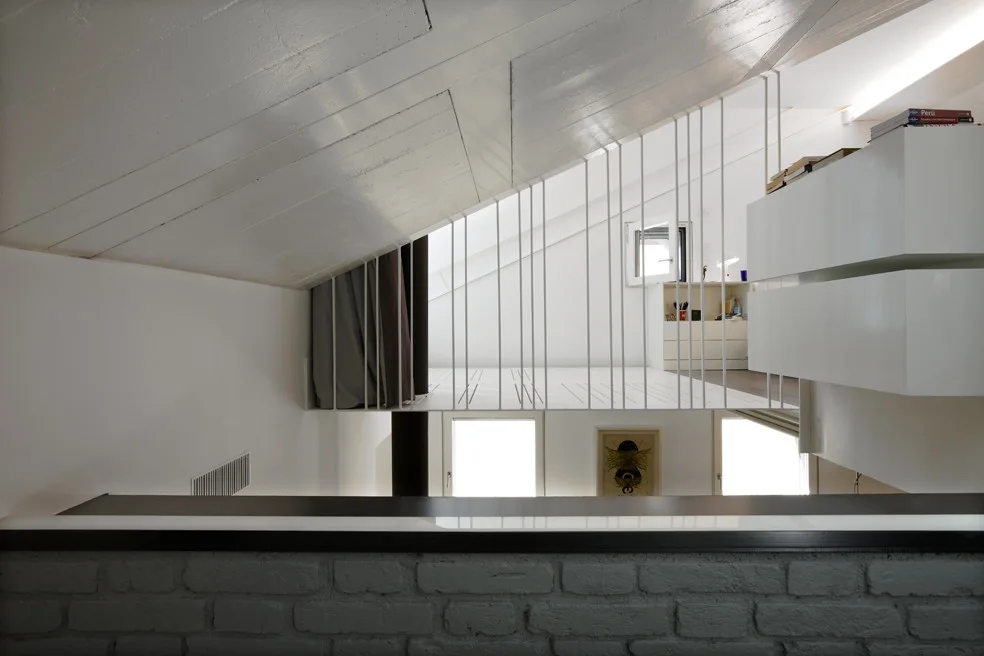 A minimalist loft mezzanine features thin white vertical metal railings under a sloped ceiling, overlooking a lower level with a painted brick wall.