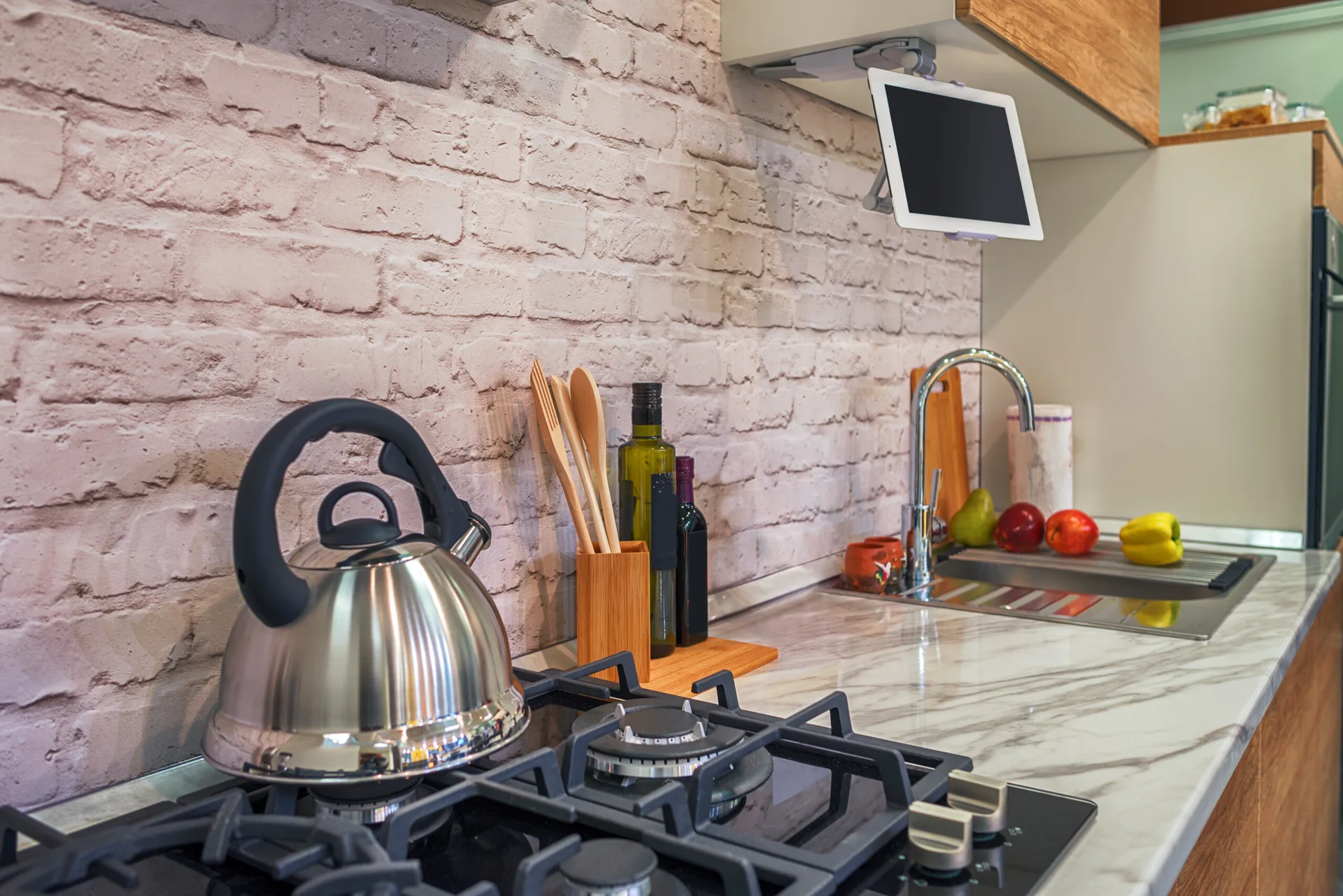 A kitchen counter features a stainless steel kettle on a gas stovetop, a white brick backsplash, and a tablet mounted beneath a wooden cabinet.