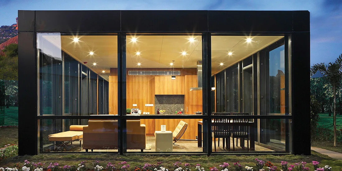 A modern glass-walled structure showcases a living space and dining area set against a warm wood-paneled kitchen wall illuminated by warm overhead lights.
