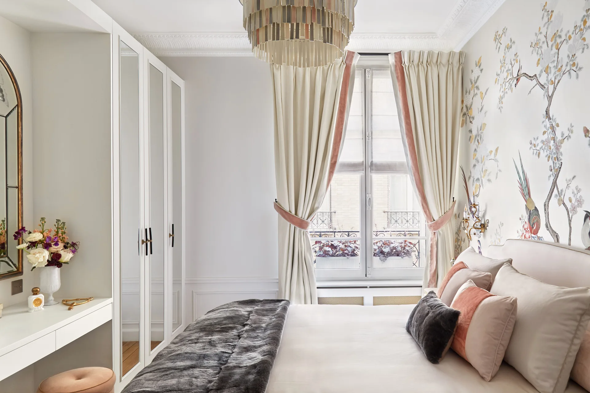 A bedroom features a built-in mirrored wardrobe, a white vanity desk, a bed with throw pillows, and soft floral chinoiserie wallpaper.