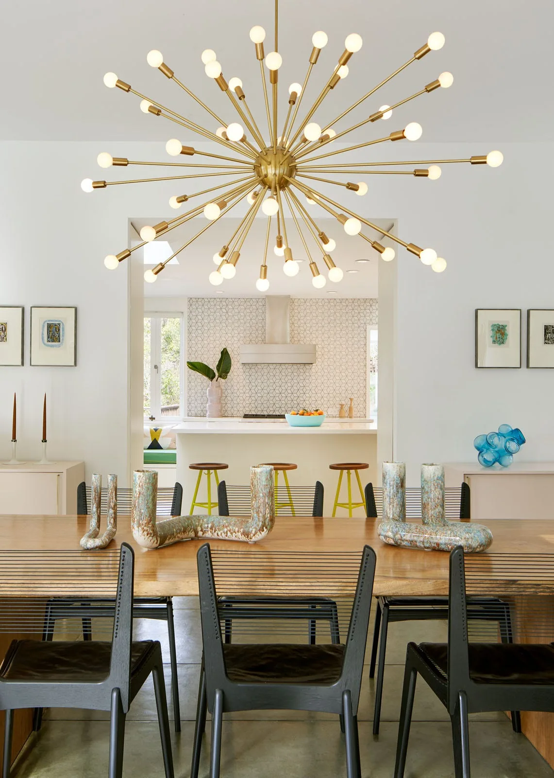 A dining room with a gold sputnik chandelier and wood table opens into a kitchen with a geometric patterned tile backsplash.