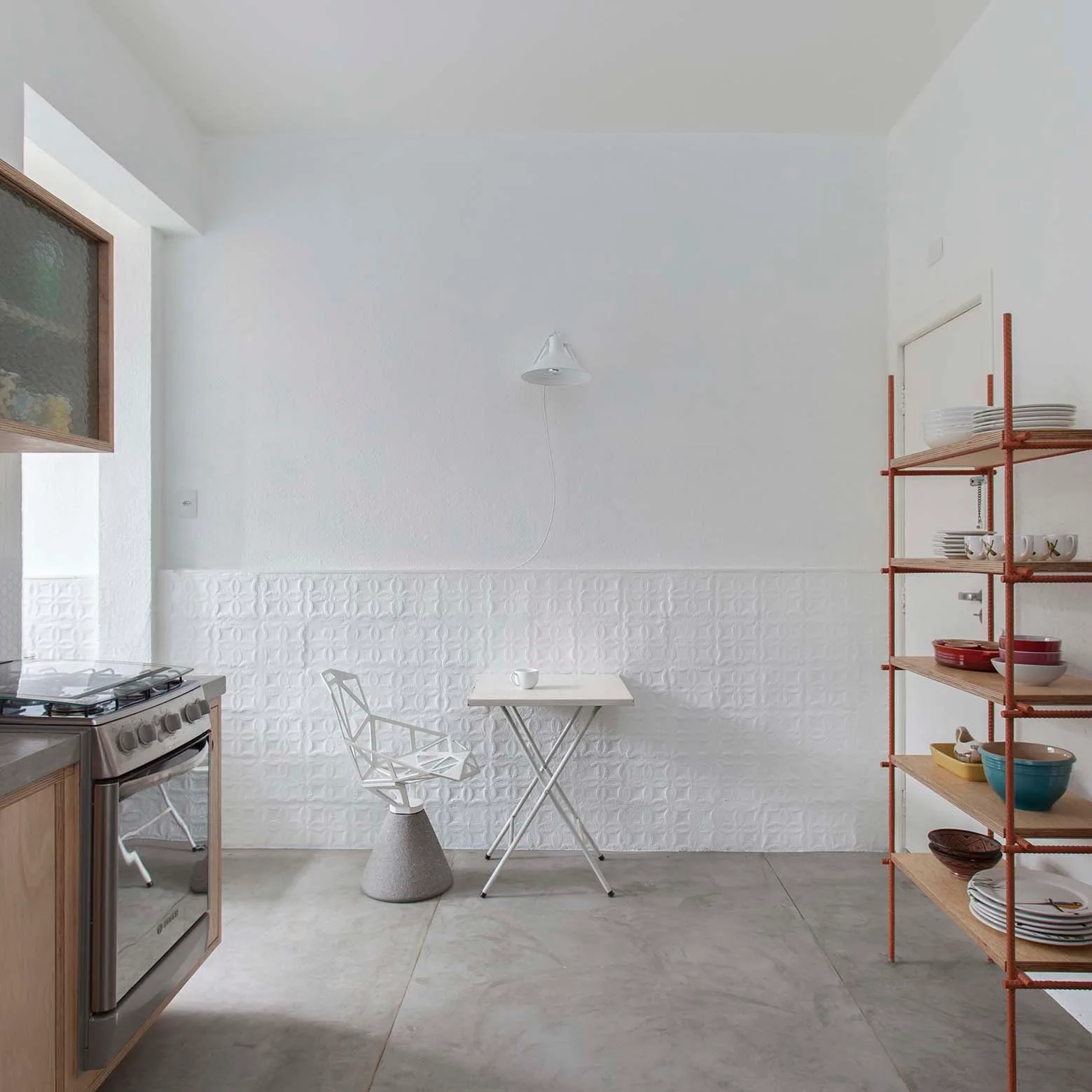 A minimalist dining nook features a small white table, a geometric metal chair, textured lower wall paneling, and an open shelving unit.