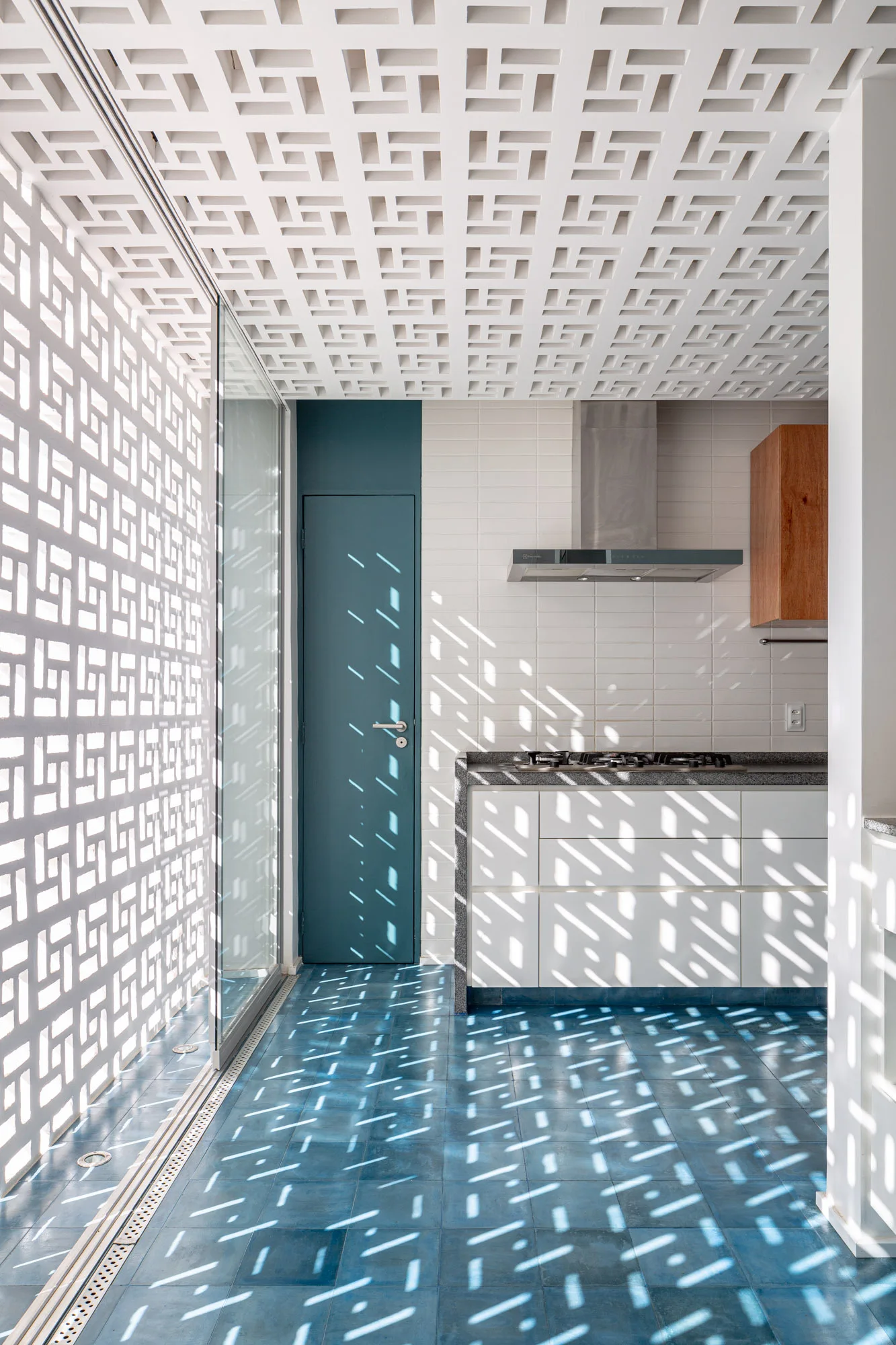 A modern kitchen with blue tile flooring, a white breeze block screen casting geometric shadows, and a teal accent door.
