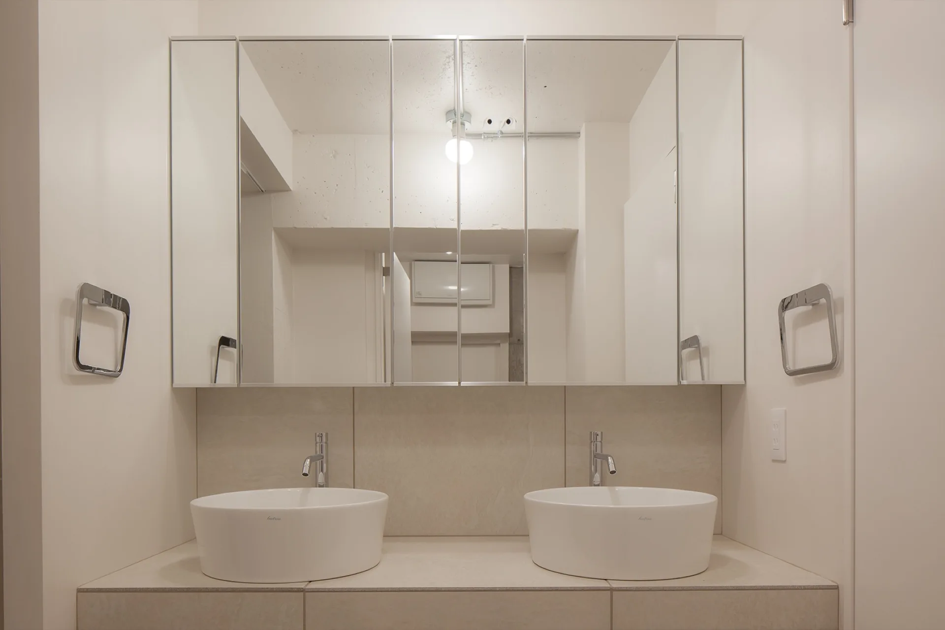 Two white ceramic vessel sinks with chrome faucets sit on a beige tiled vanity below a large multi-panel mirrored medicine cabinet.