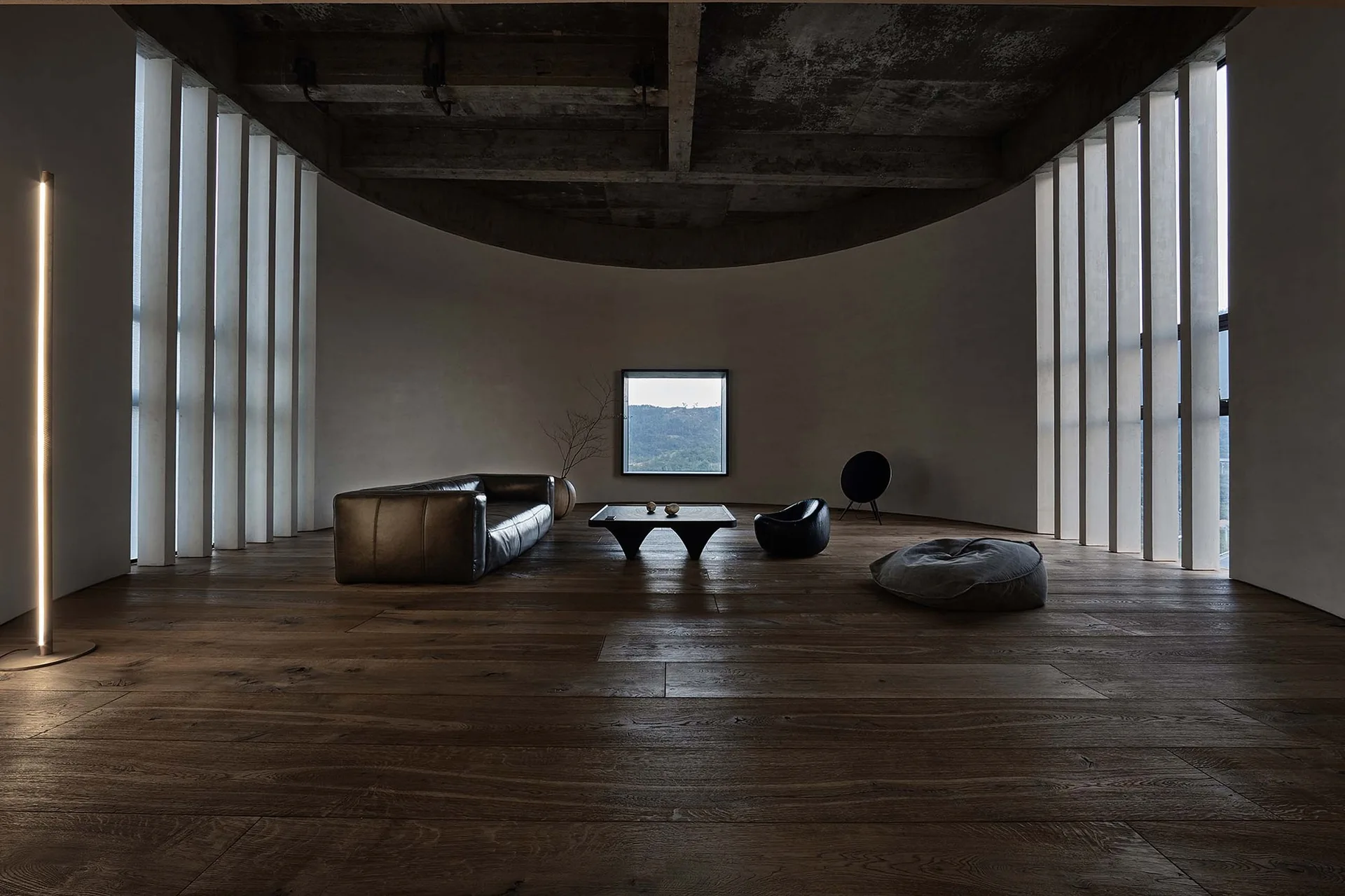 A minimalist living room with a curved wall, raw concrete ceiling, dark wood floors, and low-profile furniture.