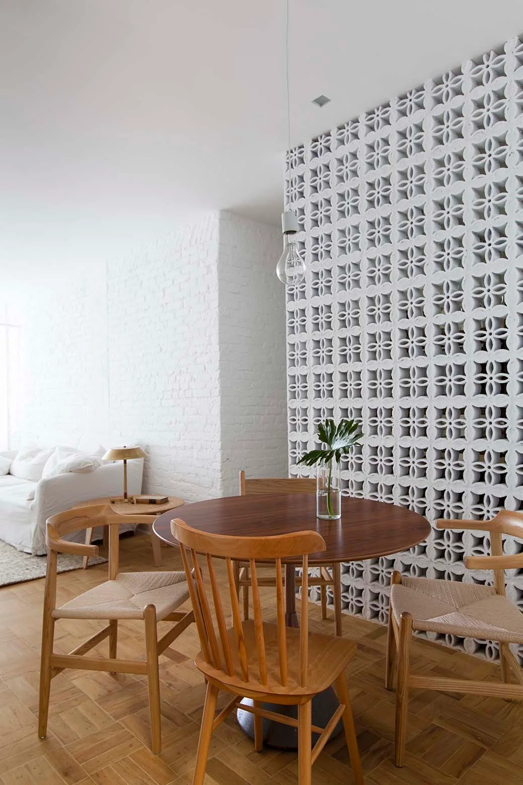 A round wooden dining table and mismatching chairs stand next to a decorative white breeze block partition and a painted brick wall.