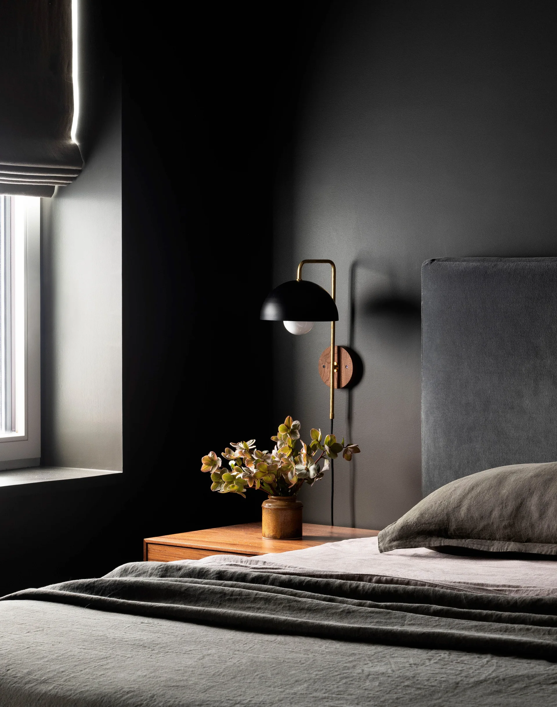 A dark charcoal bedroom corner with a wooden nightstand, a brass wall sconce, a grey headboard, and a window.