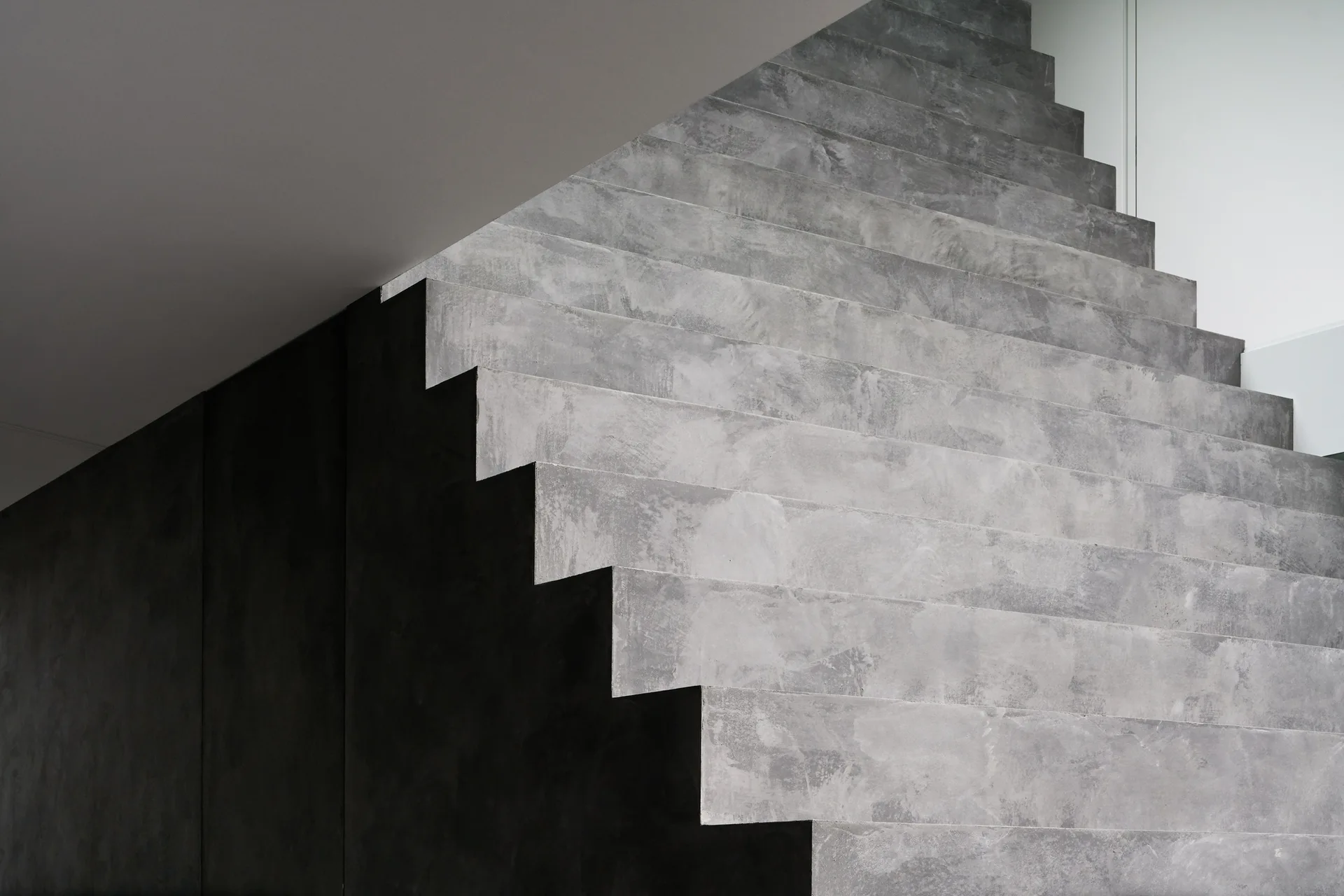 A minimalist staircase featuring textured grey concrete steps rises against a solid black wall and a white ceiling.