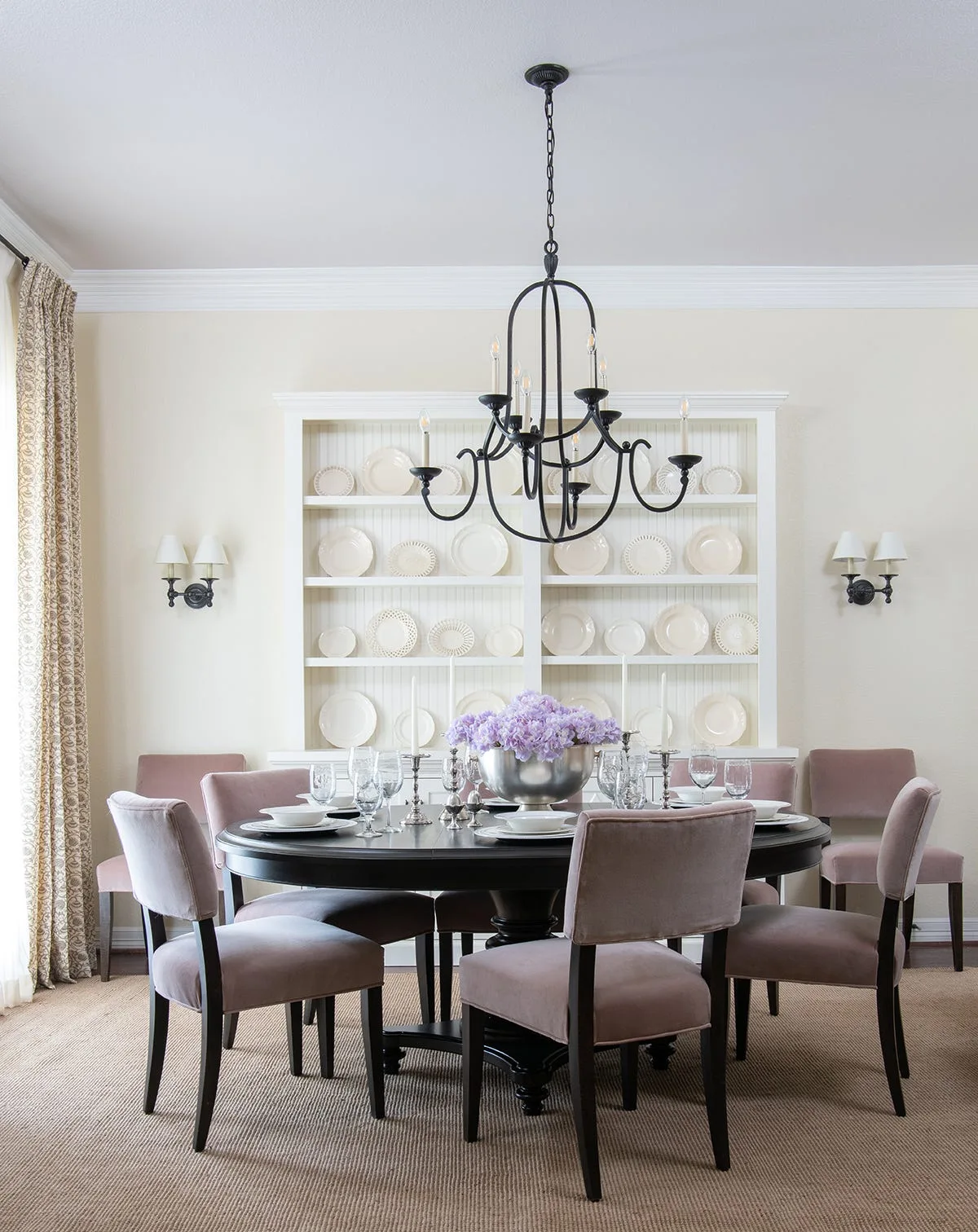 A round black dining table surrounded by upholstered dusty pink chairs sits in front of a white built-in china cabinet filled with cream dinnerware.