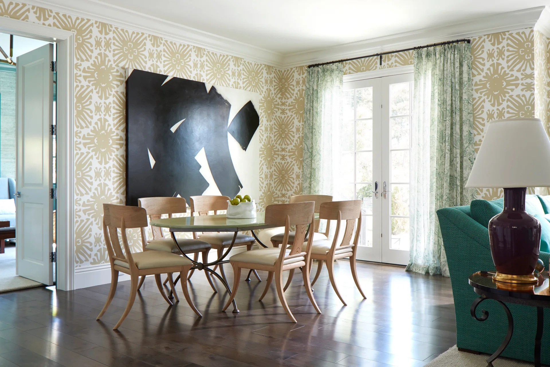 A dining room features a glass table with light wood chairs set against patterned beige wallpaper and a large black abstract painting.