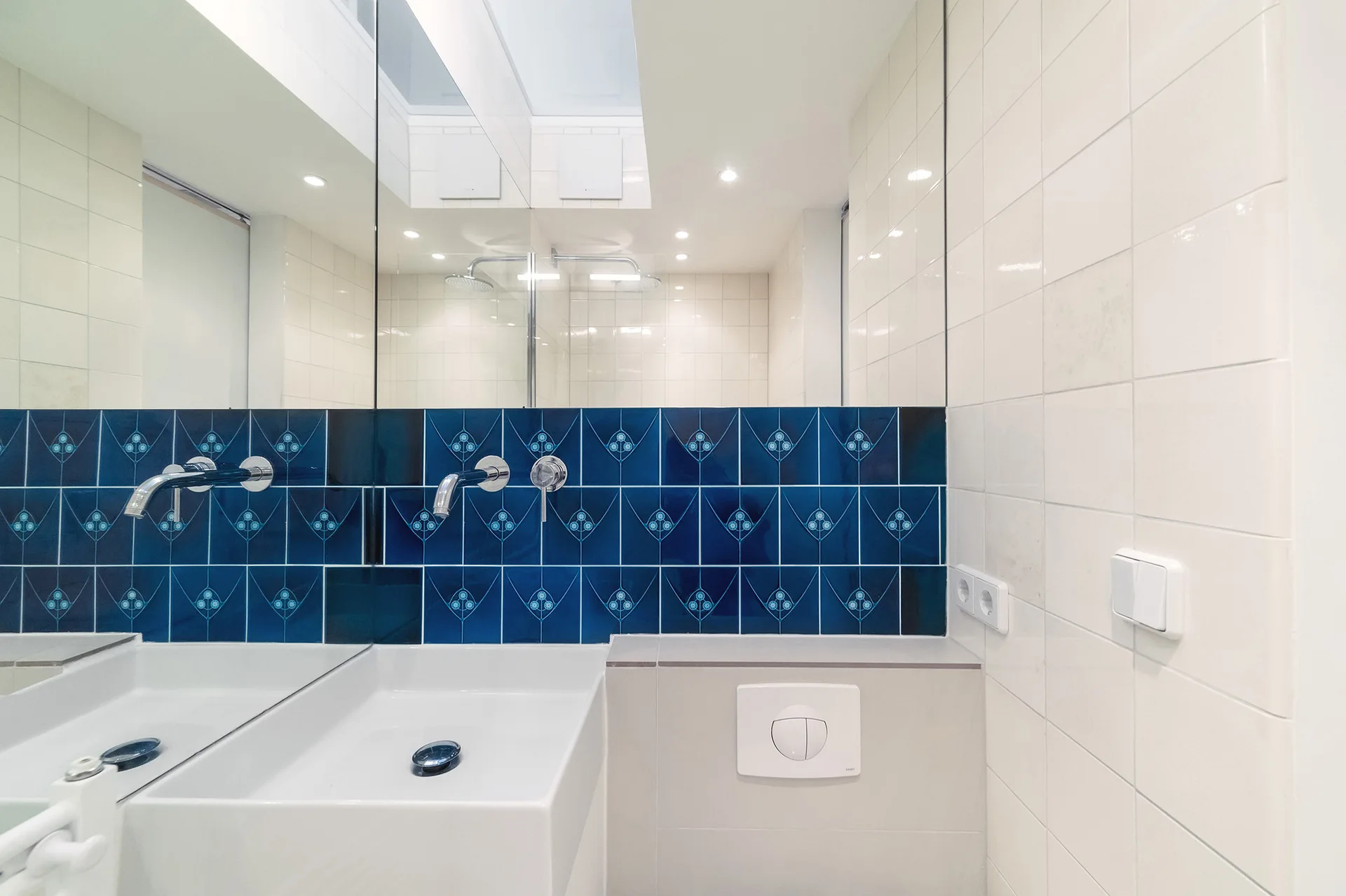 A modern bathroom features a white sink with a wall-mounted faucet set against a blue patterned tile backsplash beneath a large mirror.