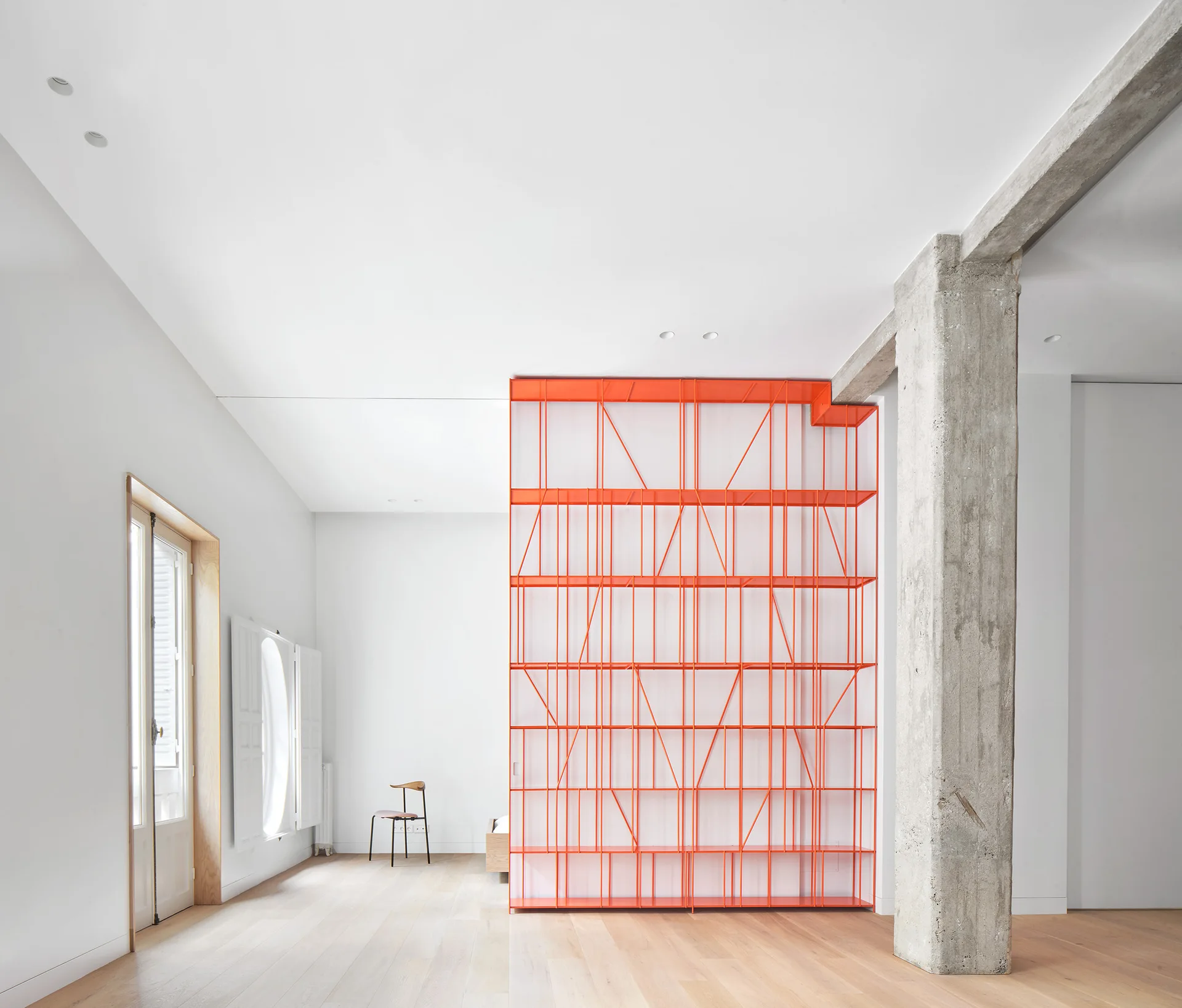 A minimalist white room features a bright orange geometric metal shelving unit, an exposed concrete column, and light wood flooring.